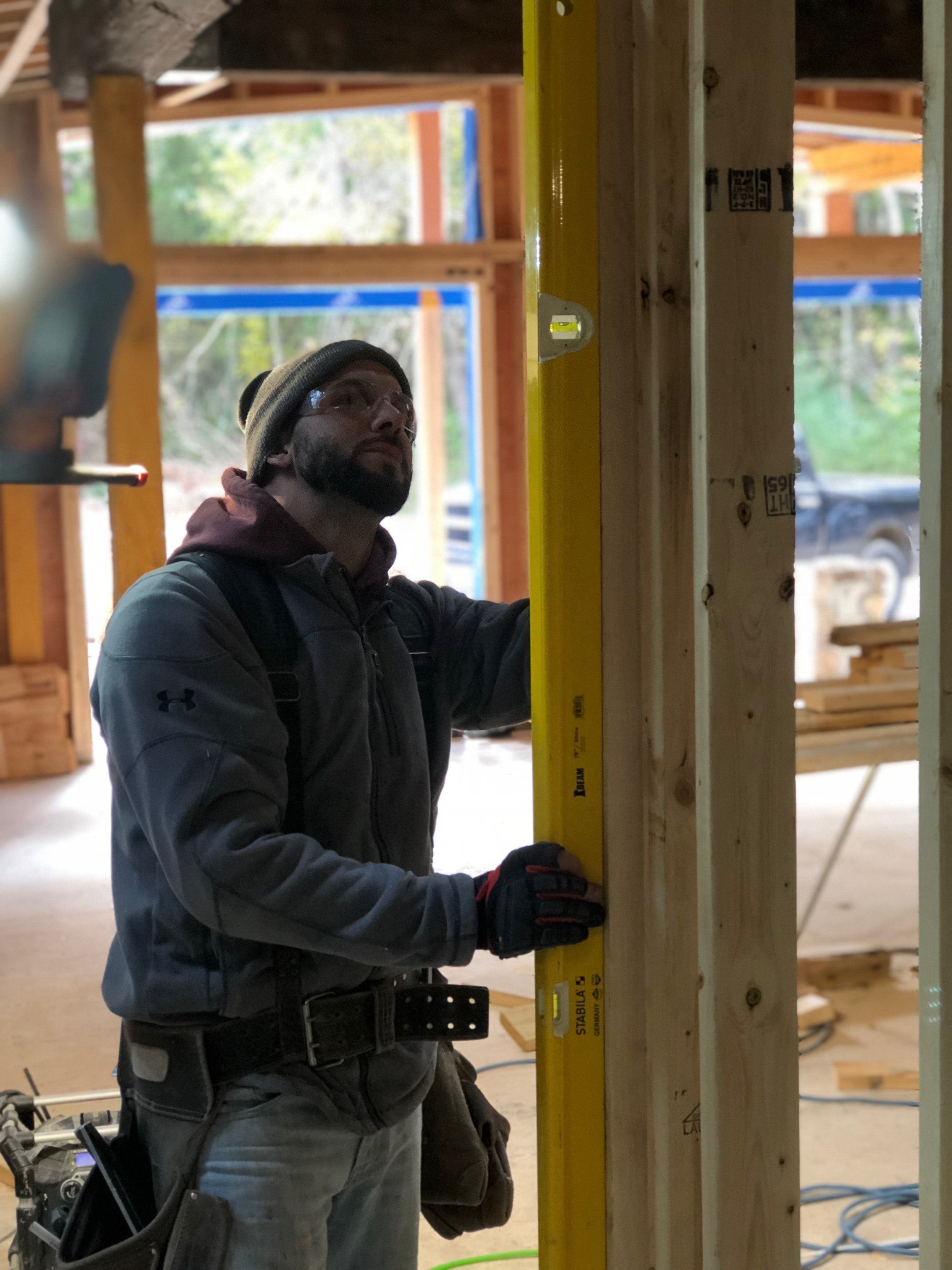 A man wearing safety glasses, a gray beanie, a dark jacket, and gloves is working with a yellow level on a wooden stud at a construction site.