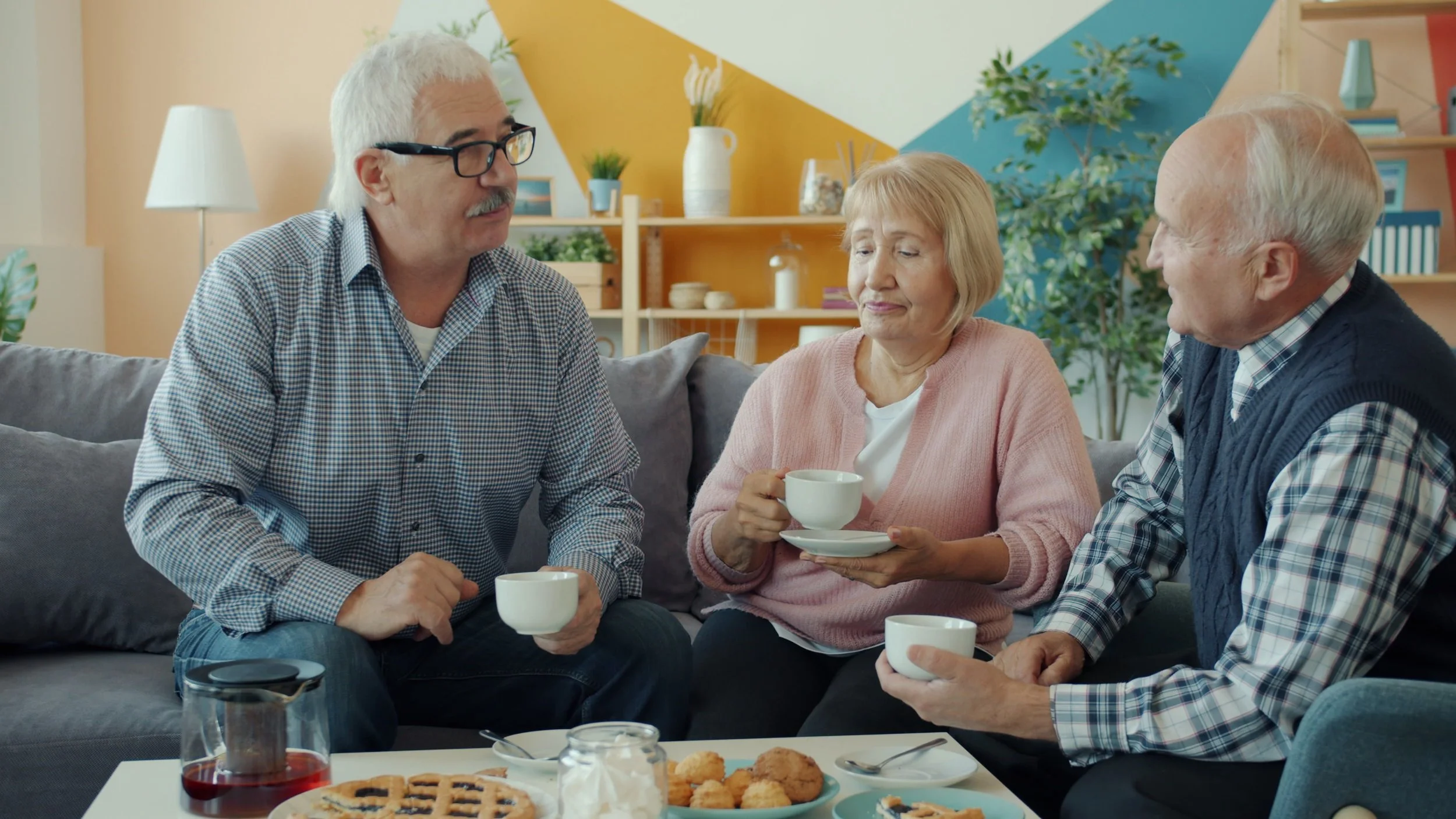 Three elderly people sitting on a couch, having a conversation with coffee cups in hand, while a table in front of them has cookies, pie, and a glass of red drink.