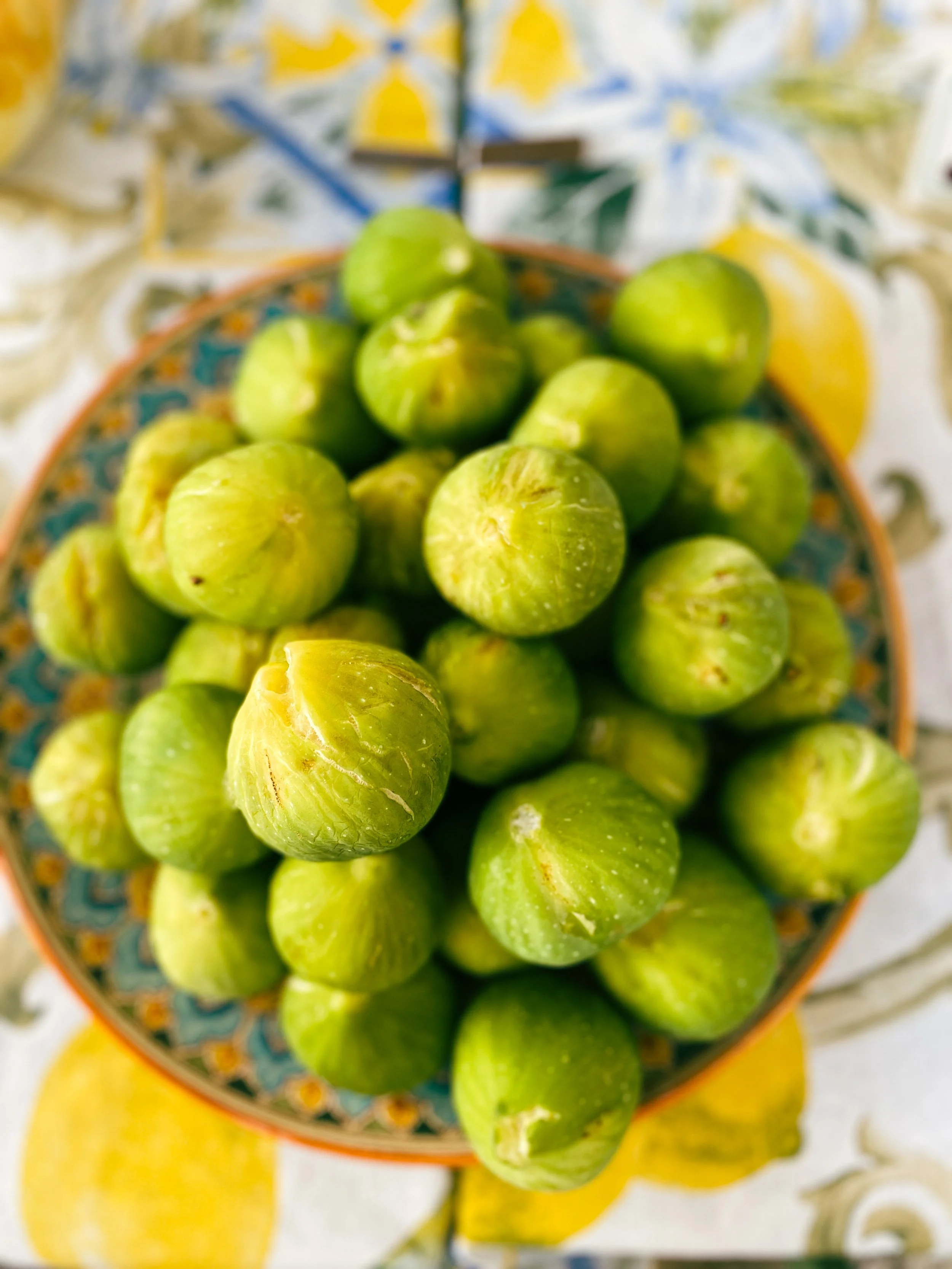 A bowl of fresh green figs on a table with lemon slices and a patterned tablecloth.