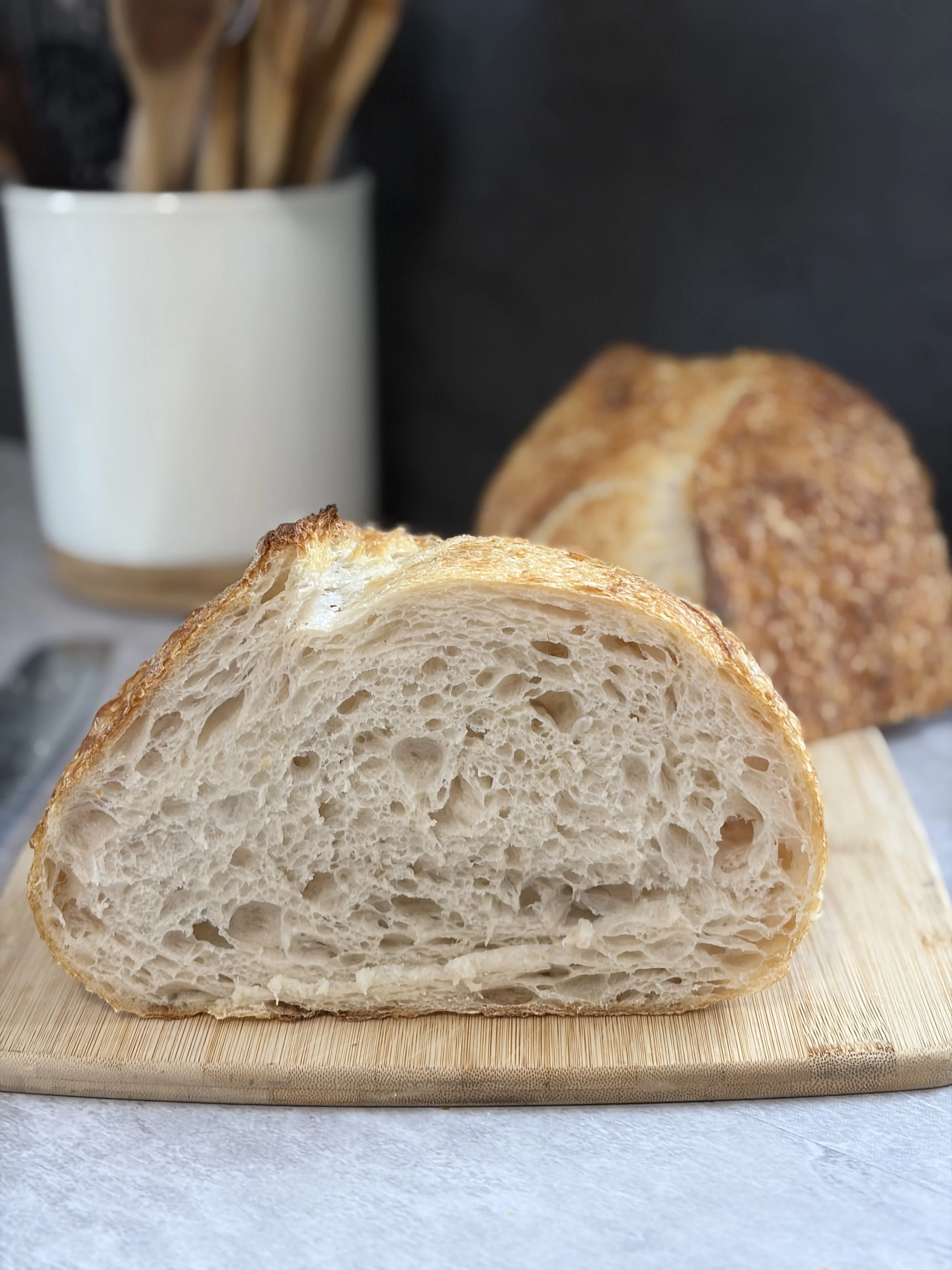 Close-up of a sliced loaf of bread on a wooden cutting board with a blurred background of a white container holding wooden utensils.