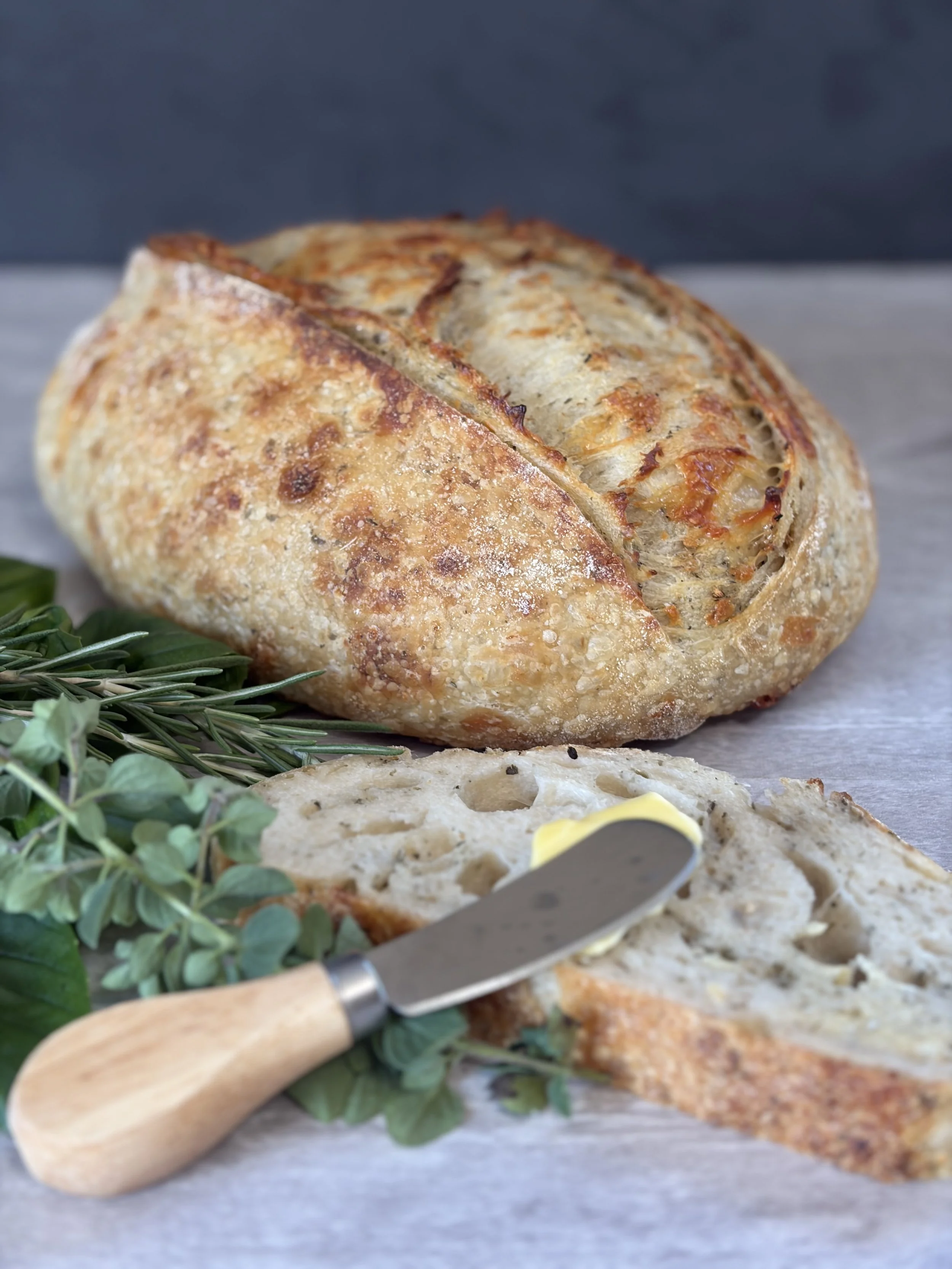 Close-up of a loaf of artisan bread with a golden, crispy crust, partially sliced, with a knife and some greenery in the foreground on a wooden surface.