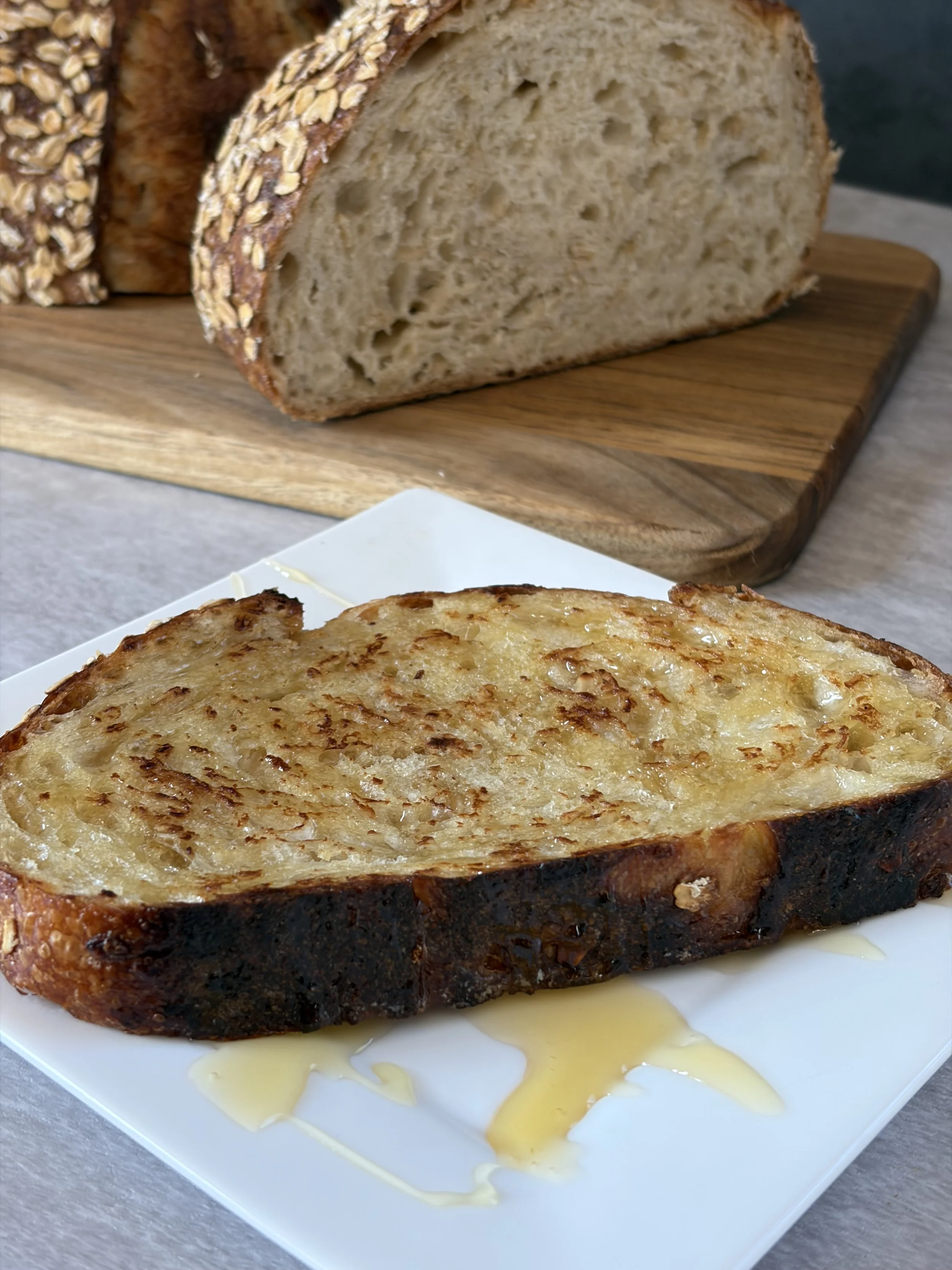 A slice of toasted bread with melted cheese on top placed on a white plate with melted butter drizzled around it. In the background, there is a loaf of bread on a wooden cutting board.