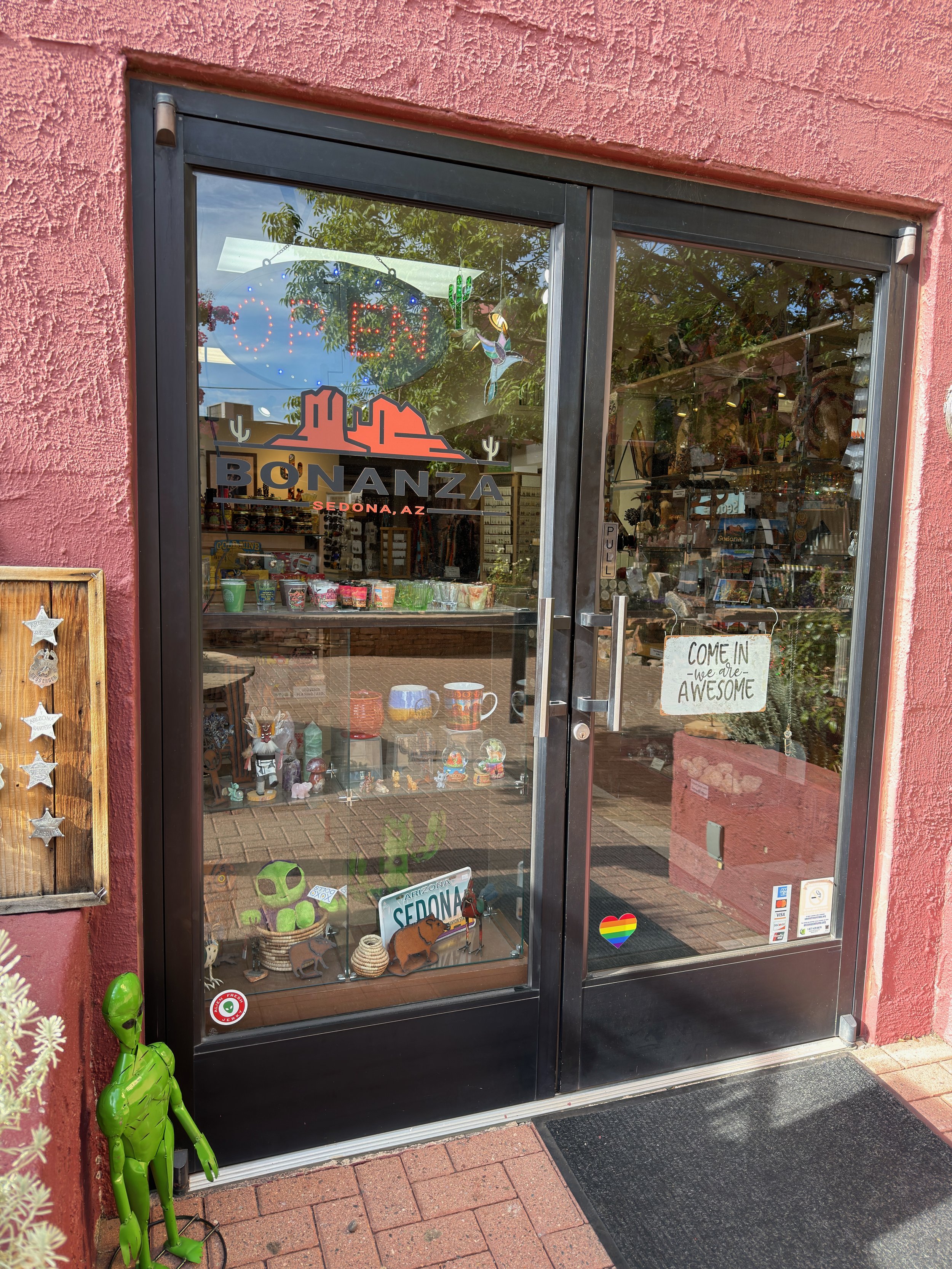 Storefront window with a sign reading "BONANZA, Sedona AZ" and various decorative items inside; outside includes a green inflatable figure resembling Hulk, a rainbow heart sticker, and a sign that says "Come in - we are awesome."