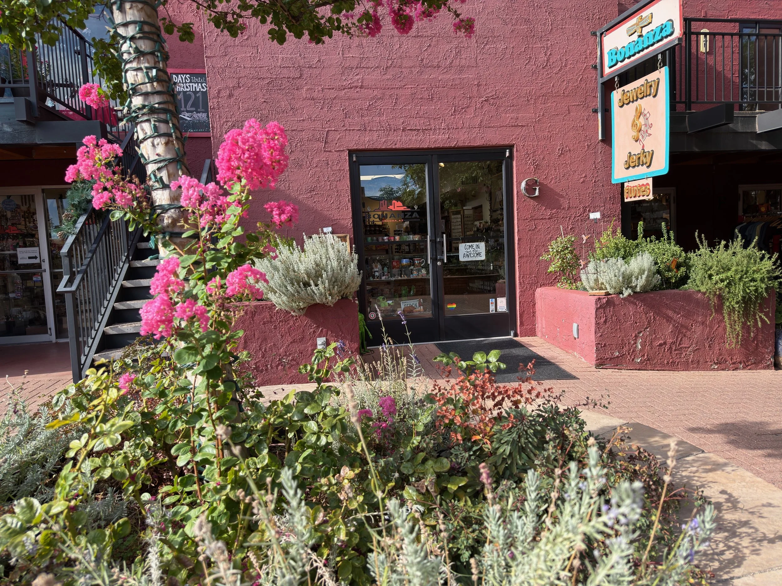 The exterior of a storefront with pink textured walls and colorful signs advertising jewelry, Jerky, and fudge. The entrance has a glass door with a sign that says 'Come in, we are awesome.' There are pink flowers and green plants in front of the entrance and a staircase leading to an upper level on the left.