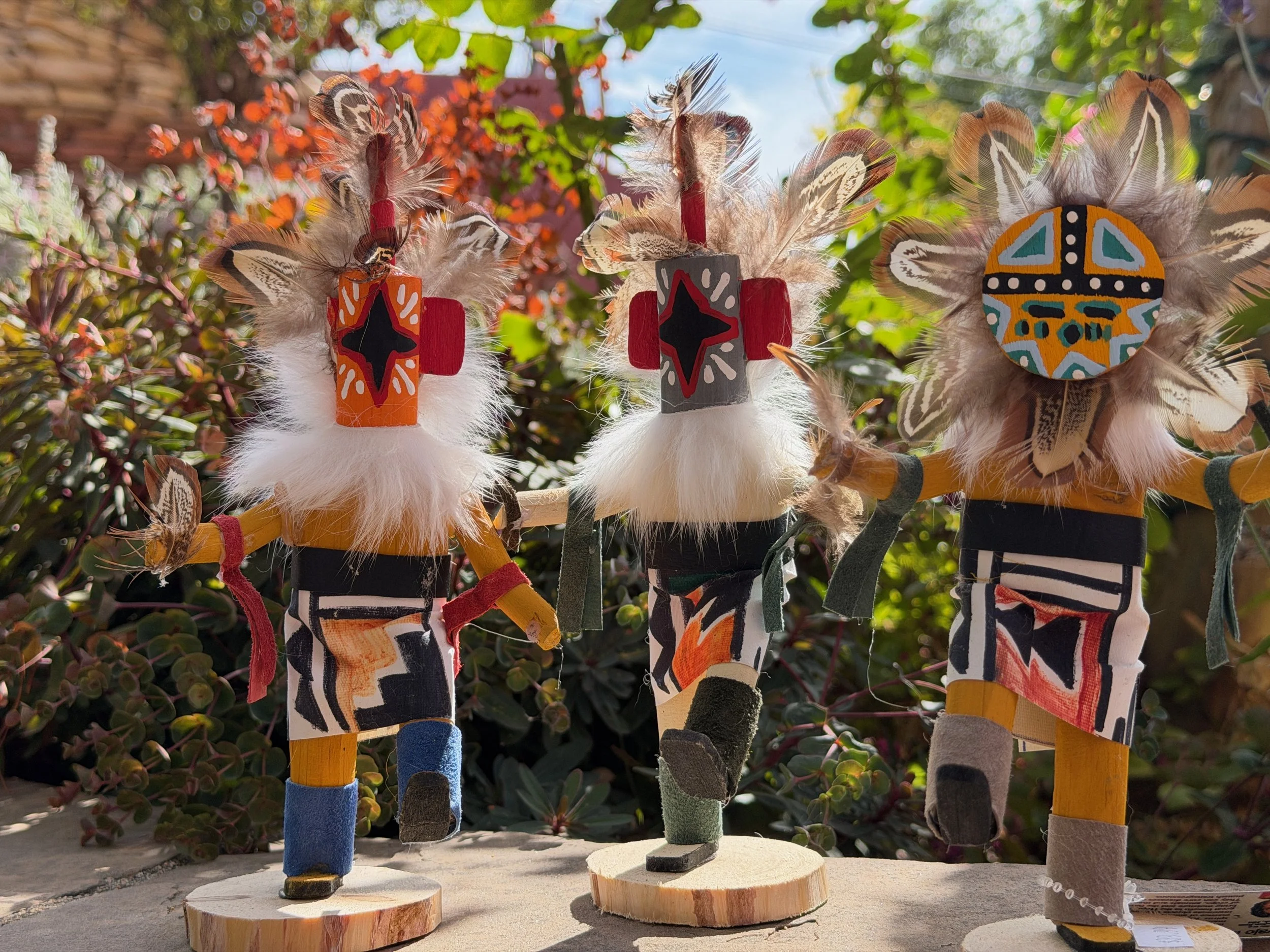Three decorative figurines of Native American characters with feathers and painted faces, standing on wooden bases outdoors with greenery in the background.