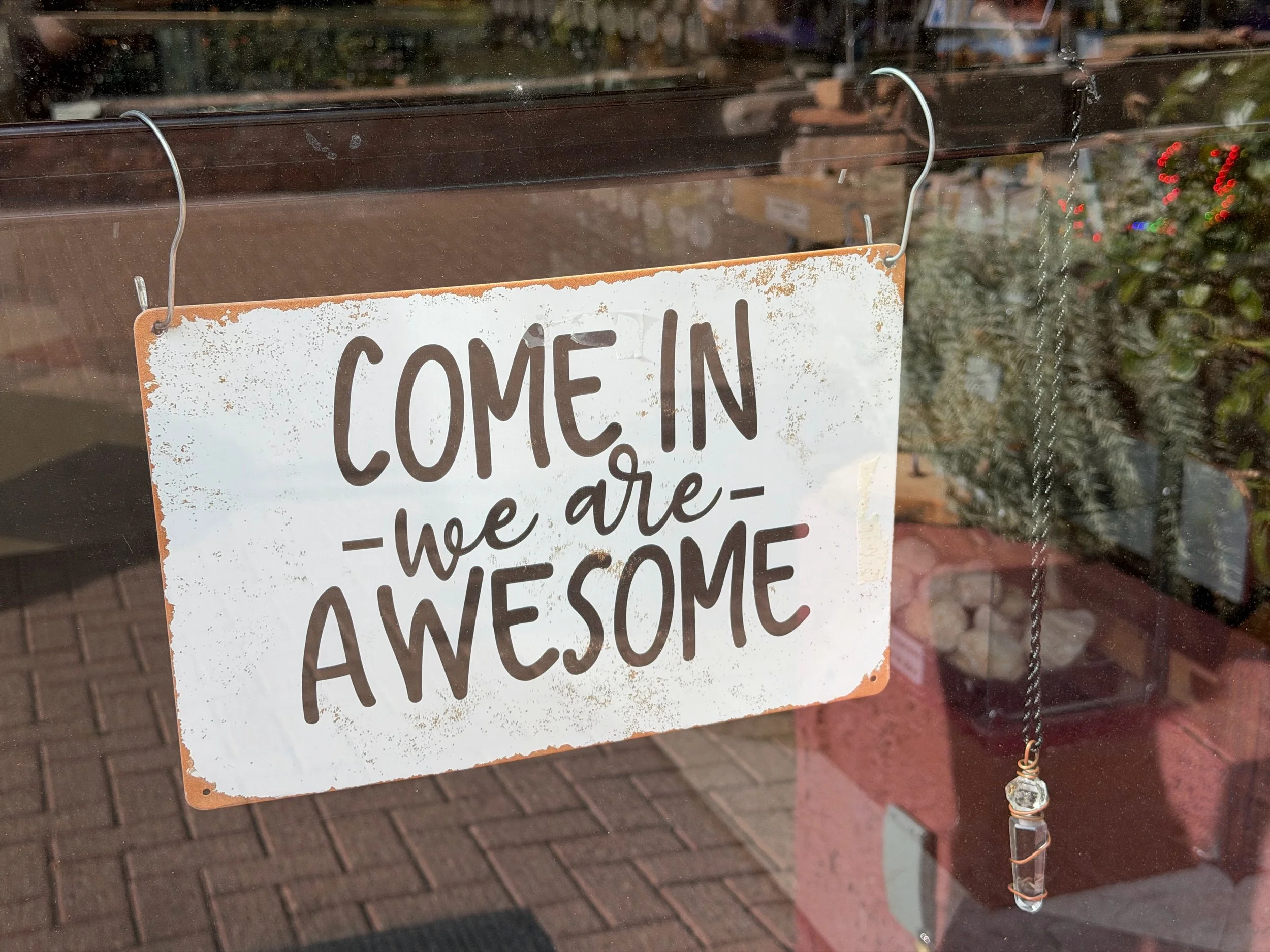 A close-up view of a glass door with a sign that says 'Come in, we are awesome,' hanging from two metal hooks. There are reflections on the glass and some blurry background elements visible.
