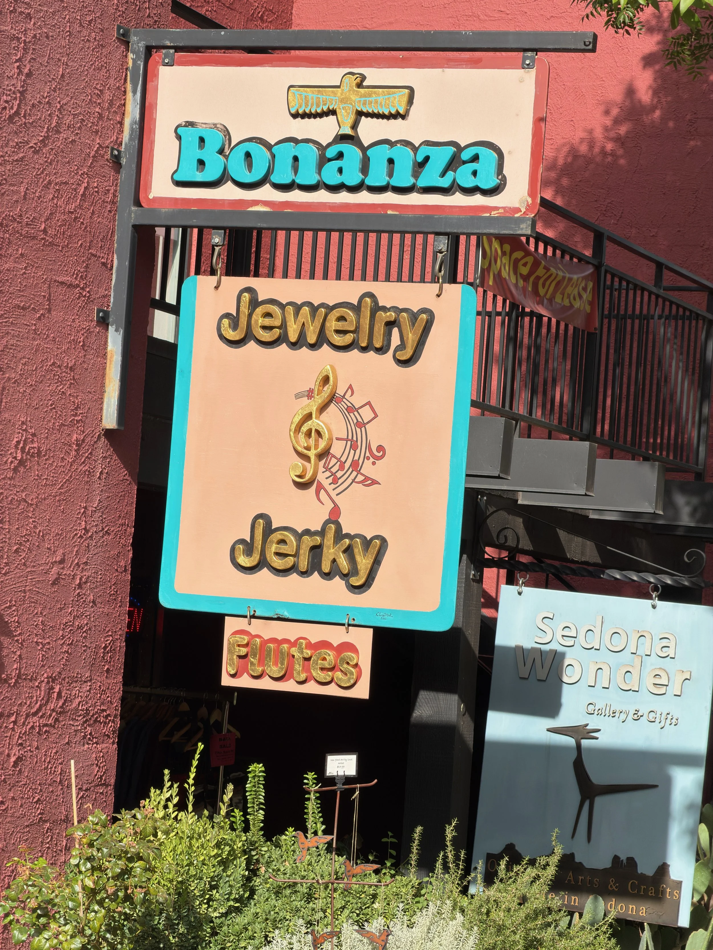 Storefront sign for Bonanza Jewelry with a separate sign for Flutes, featuring a treble clef, surrounded by plants, against a red textured wall. Other signs partially visible around.
