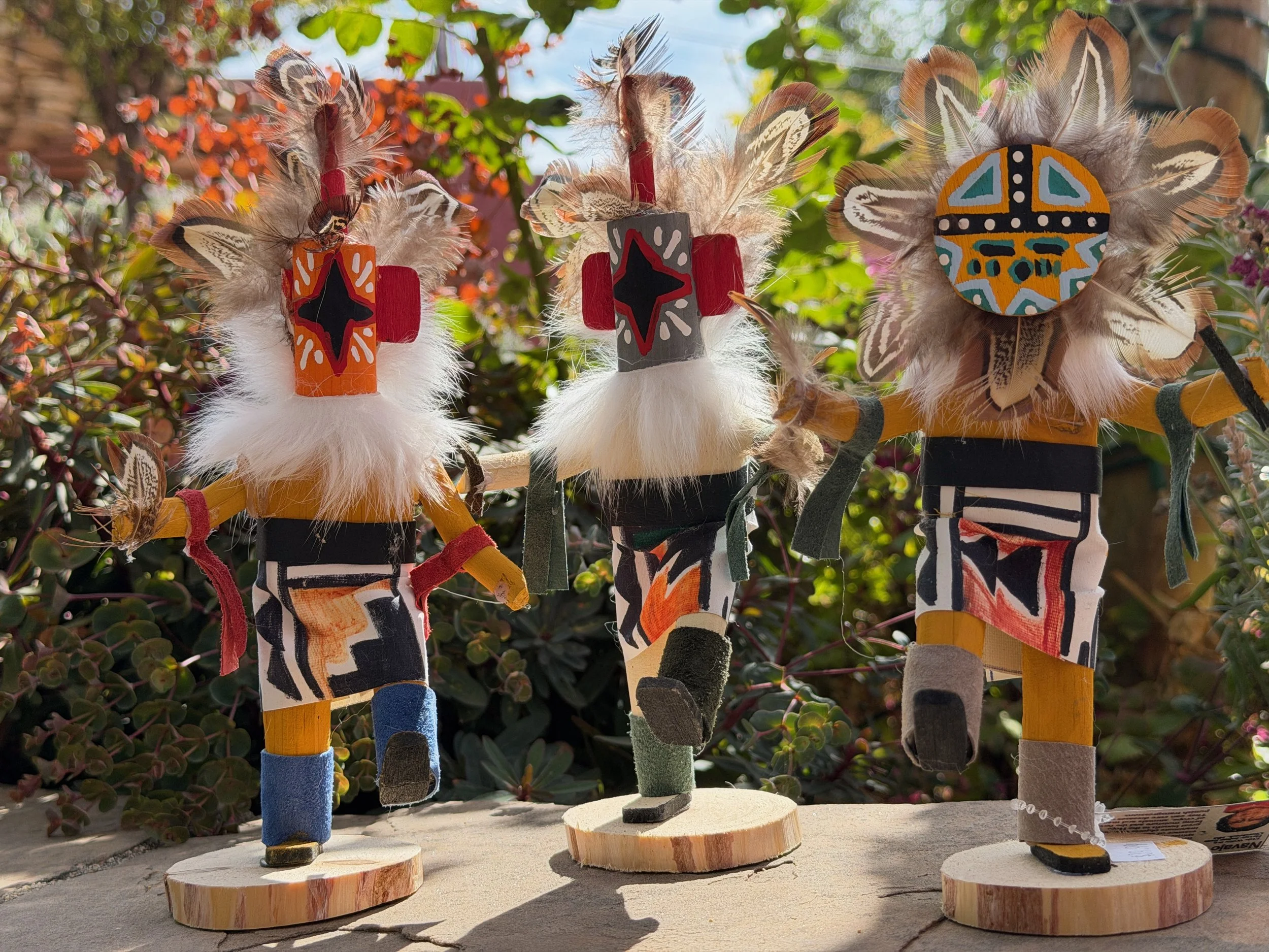 Three decorative figurines made of wood, feathers, and painted designs are standing on wooden bases outdoors, surrounded by plants and sunlight.