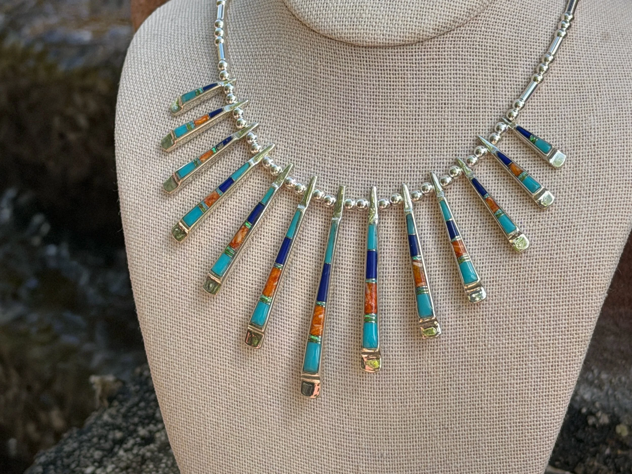 A silver statement necklace with colorful inlays of turquoise, orange, and blue, worn on a beige fabric surface.