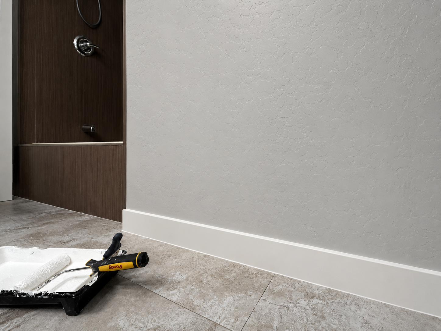 Part of a room with gray textured wall, baseboard, and tile floor. There is a dark tile built-in tub/shower combo, on the wall with plumbing fixtures and a metal handle. A painting roller and paint tray with white paint are on the floor.