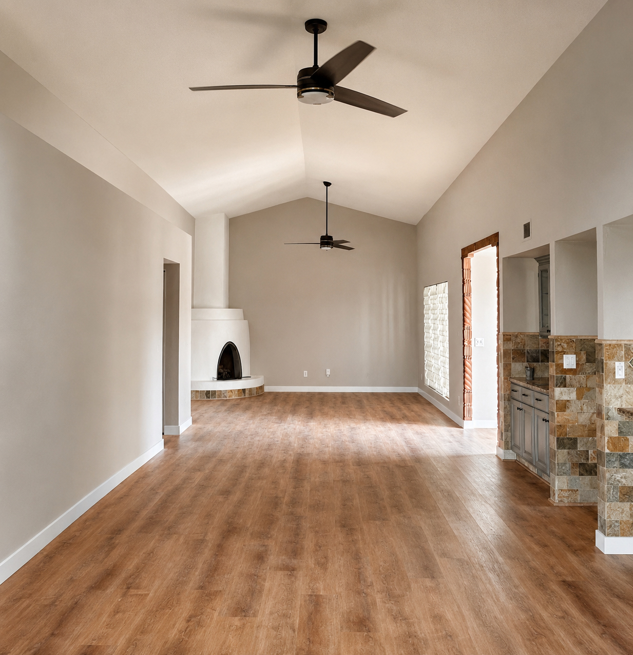 Empty living room with wood flooring, high vaulted ceiling, two ceiling fans, fireplace with a curved white mantel, brick accents around an open doorway, and a small built-in bar area with stone tiles.
