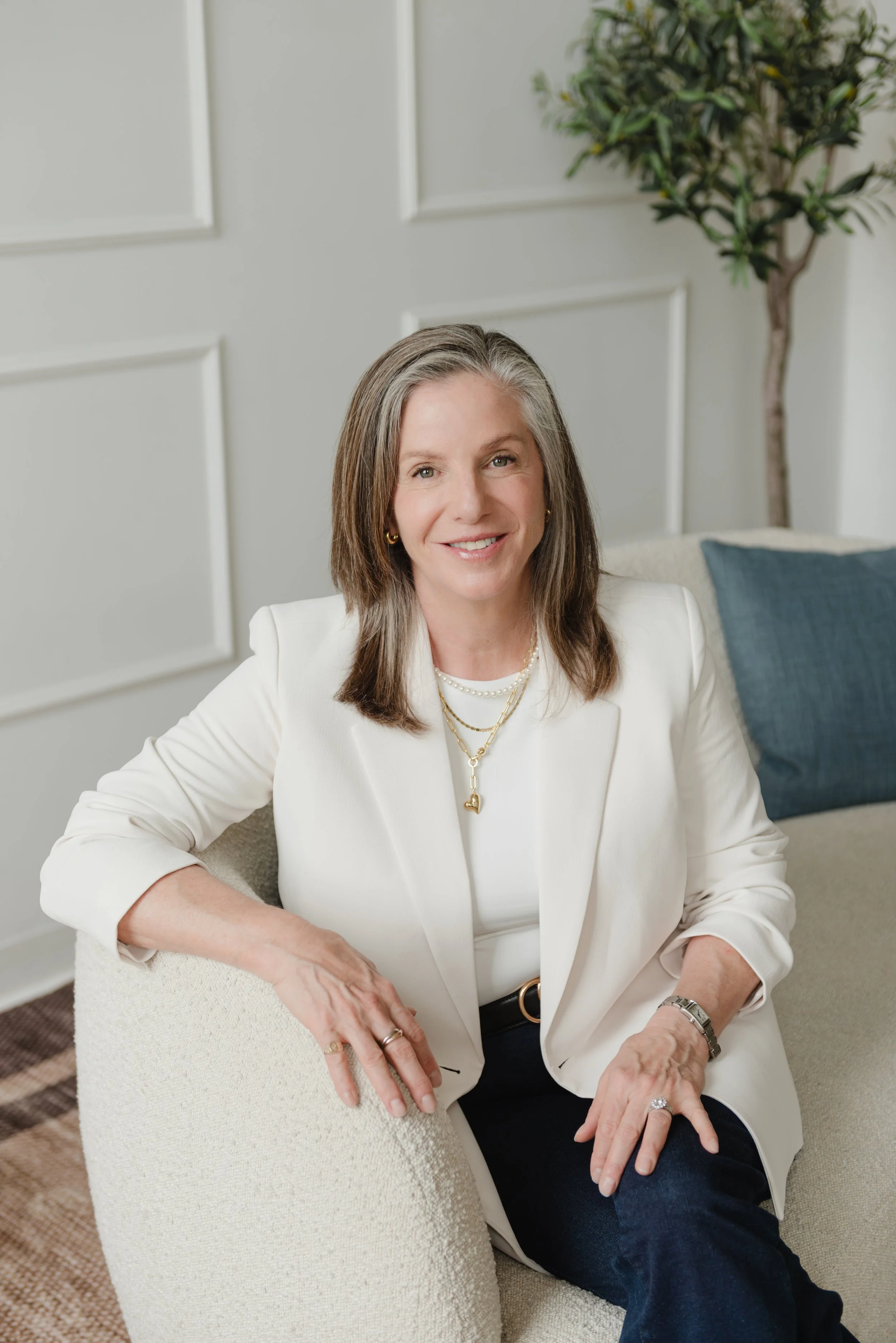 Wendy Shami wearing a white blazer, sitting on a beige chair, smiling at the camera. She is accessorized with gold and pearl necklaces, rings, and a watch. The background includes a white wall with decorative molding and a green potted plant.