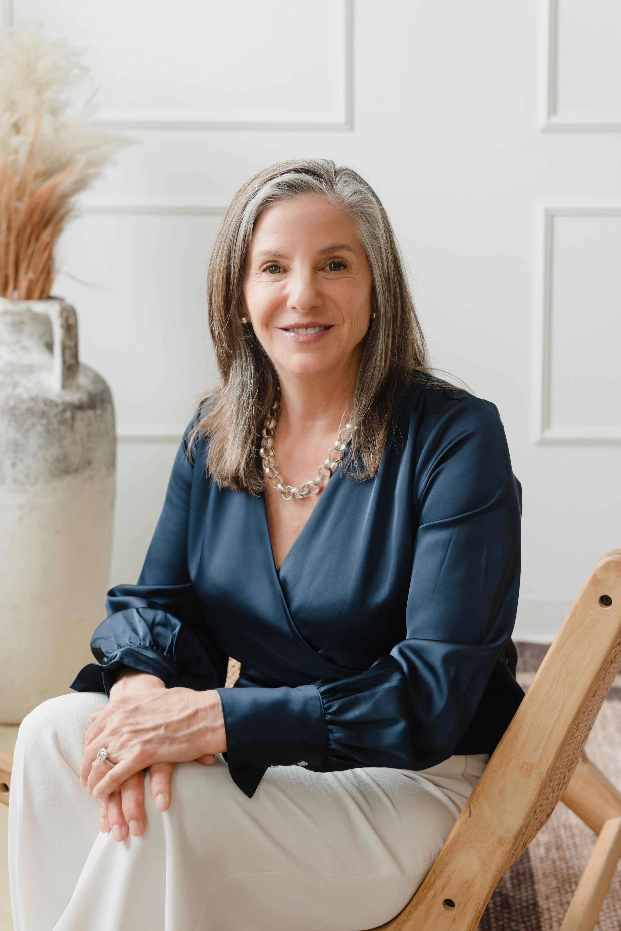 Wendy Shami headshot. Sitting on a wooden chair, wearing a navy blouse and white pants, smiling, with a large textured ceramic vase holding dried plants beside her in a bright, modern room.