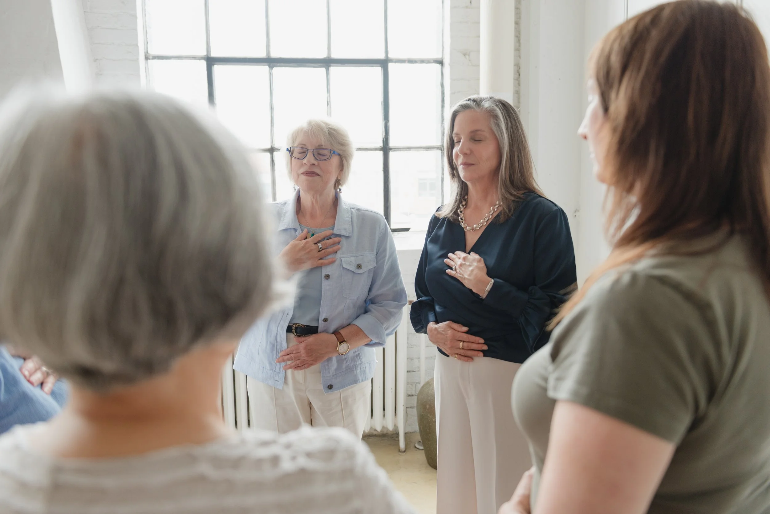 A group of women standing in a circle with their hands on their chests in a somatic experience.