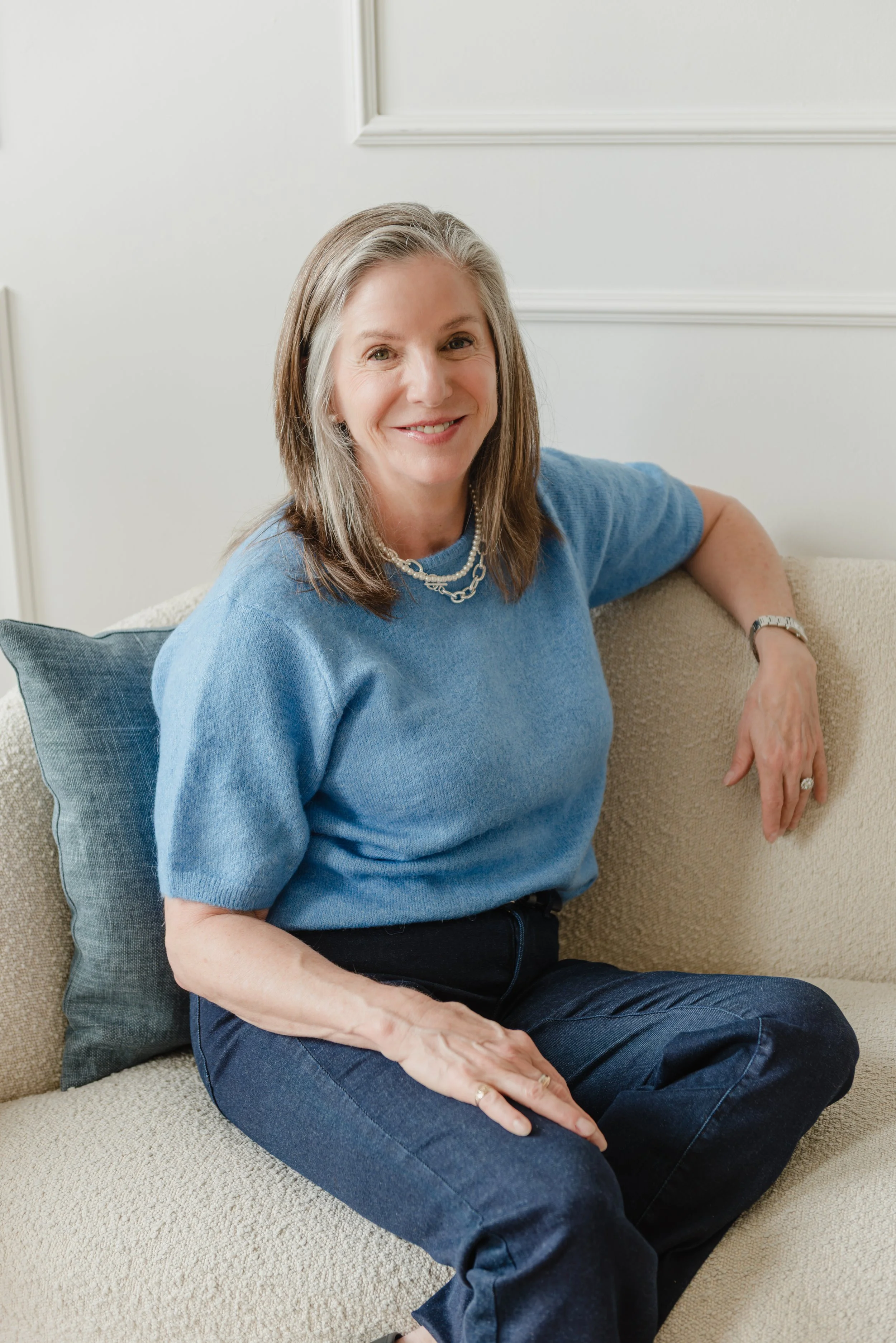 Wendy Shami wearing a blue sweater, black jeans, and a pearl necklace, sitting on a beige sofa with a dark gray pillow, in a room with white walls and decorative molding.