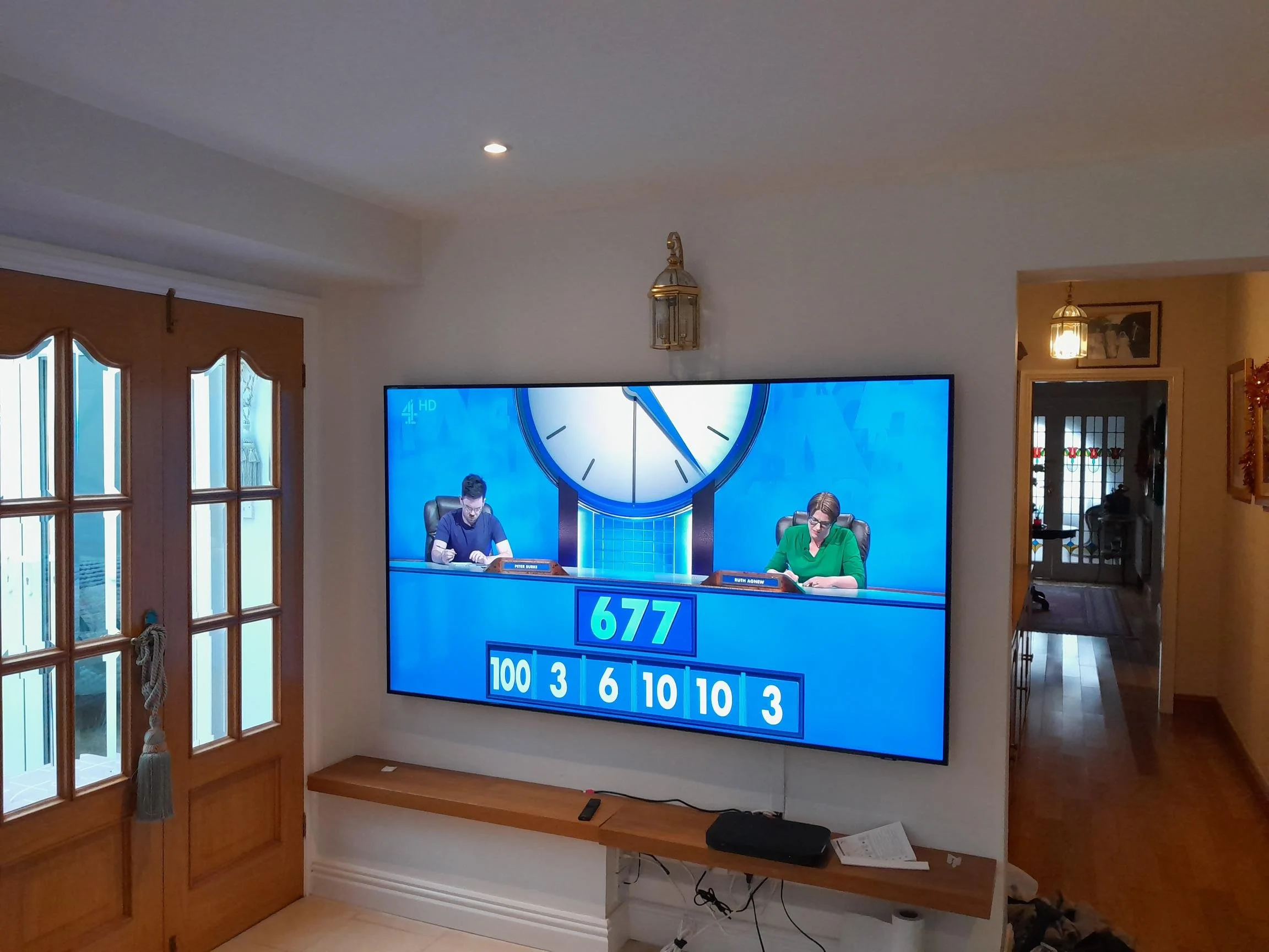 A Wall mounted large screen television displaying a quiz game, with two contestants seated at a table, shown on a wall in a living room. The room has wooden furniture and glass-paneled doors.