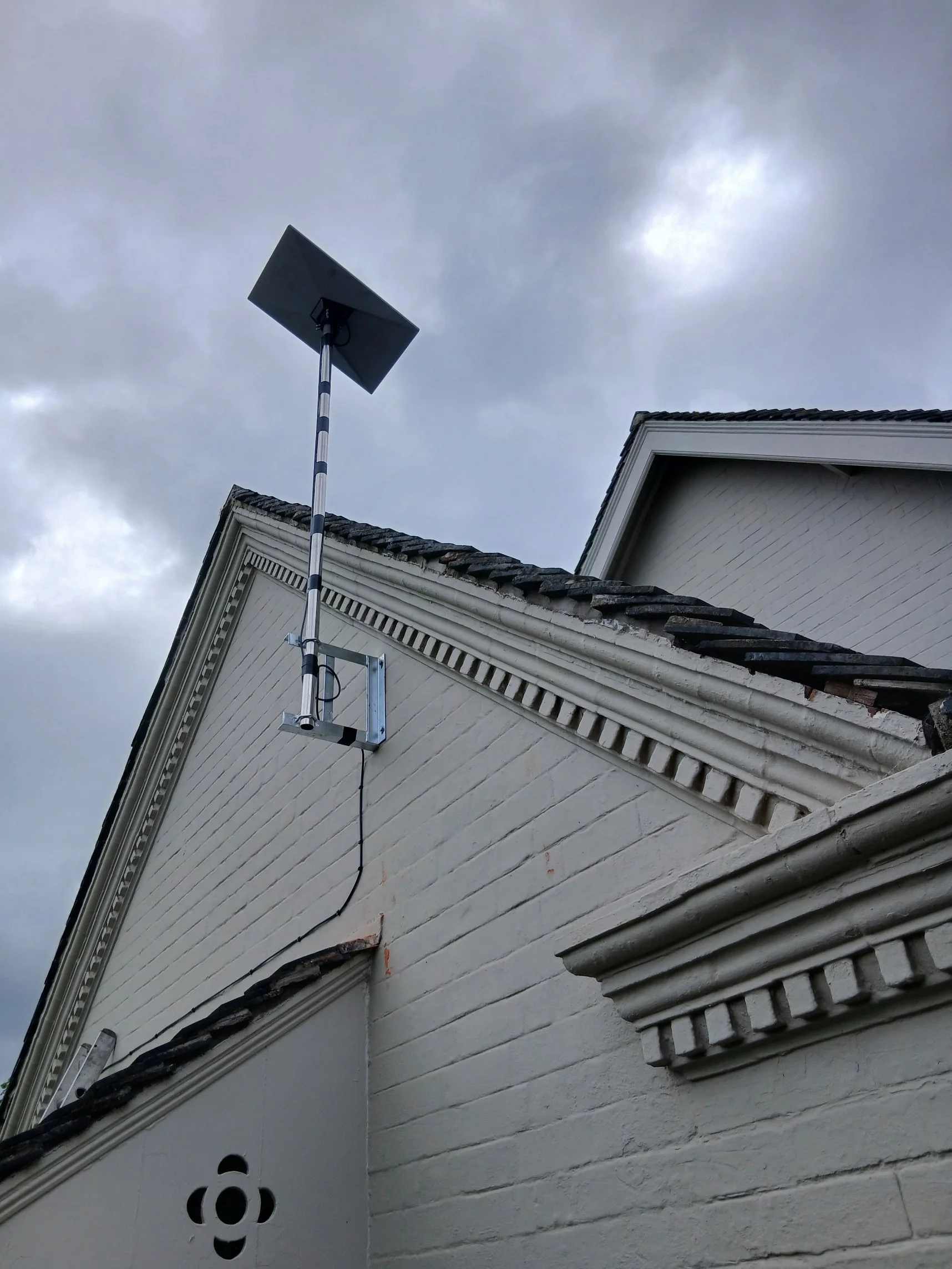 Close-up of the upper corner of a white house with a sloped roof, a black and white striped antenna mounted on the wall, and the sky with gray clouds overhead.