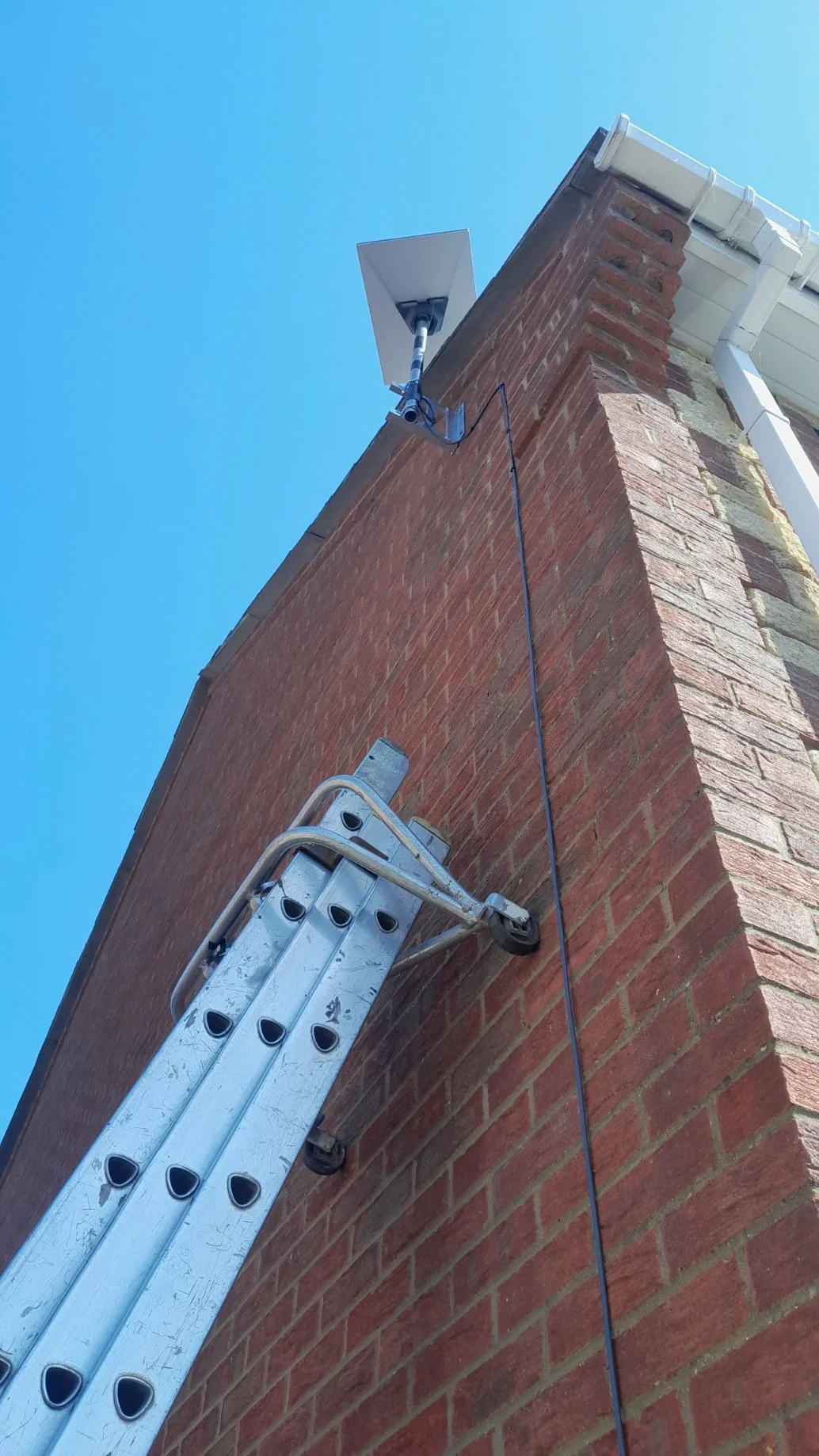Ladder leaning against a brick chimney, with a security camera mounted on the chimney, and a gray cable running down the brick wall, under a clear blue sky.