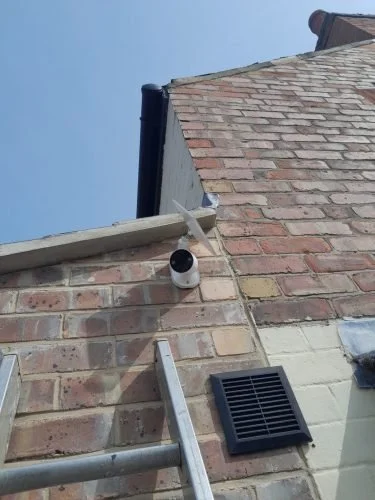 Close-up of a brick building corner with security camera, ladder, and ventilation grille against a close blue sky.