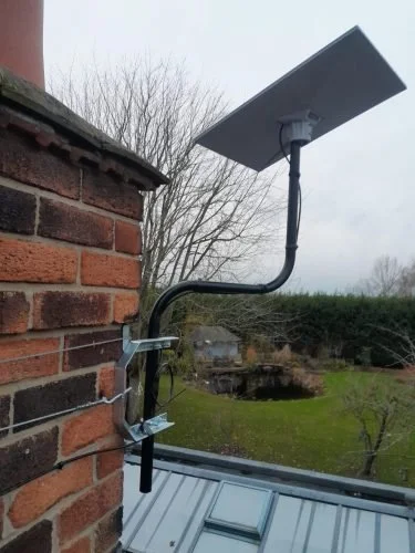 A satellite dish mounted on a brick wall with cables connected to it, set in a backyard with trees and a cloudy sky in the background.