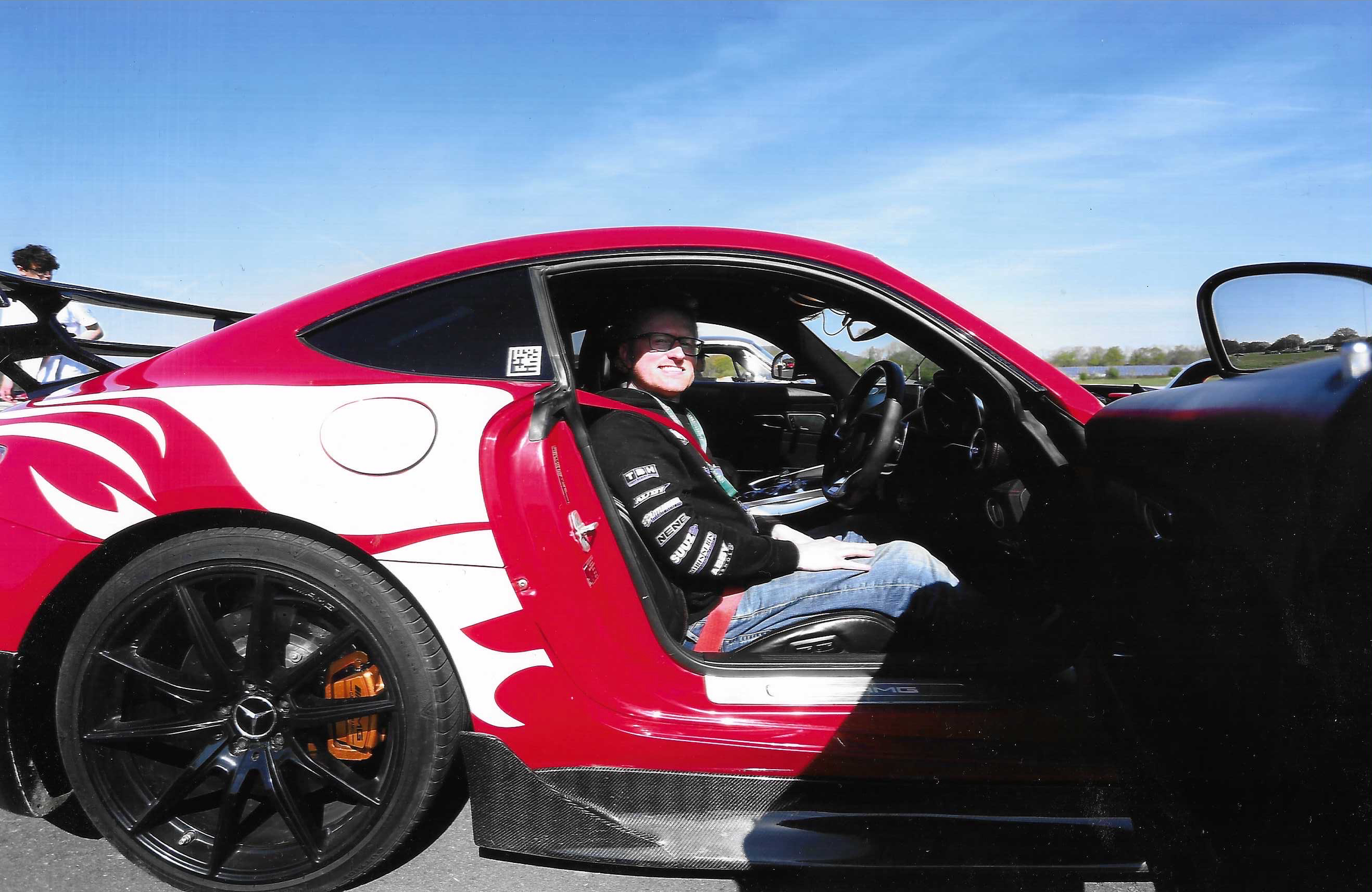 A red and white sports car with a person smiling in the driver's seat, wearing glasses and a racing suit with sponsorship logos, parked on an outdoor road under a clear blue sky.