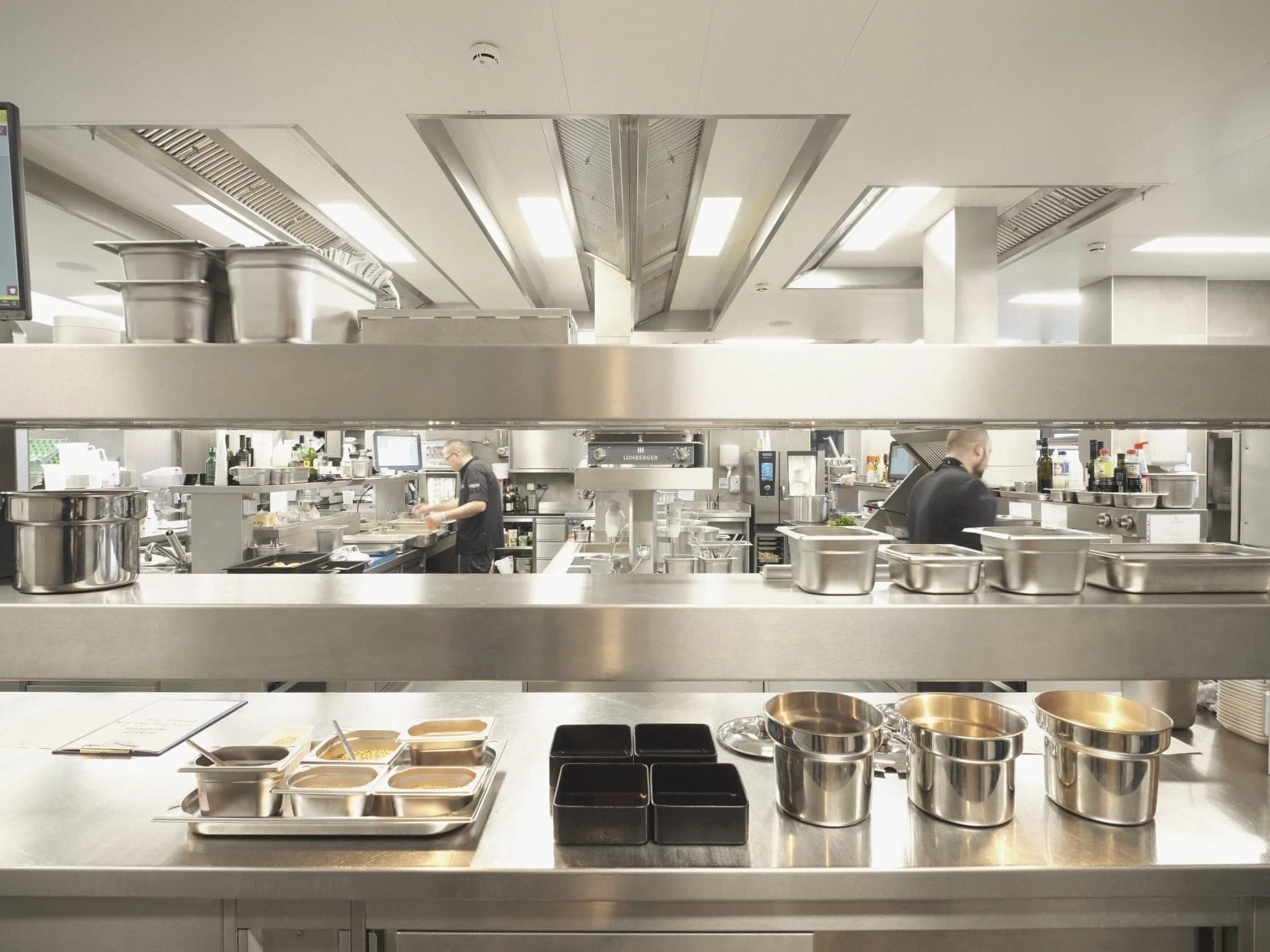 Gastro-Küche von Lohberger Küchentechnik mit Edelstahl-Arbeitsflächen, Töpfen, Schüsseln und Küchenutensilien, Küchenpersonal arbeitet an den Stationen