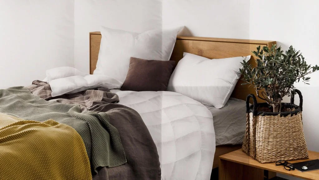 Ein Bett mit weißen Daunenkissen von Sanders-Kauffmann, einem braunen Federkissen, einer Daunendecke von Sanders- Kauffmann und einer gelb-grünen Tagesdecke.