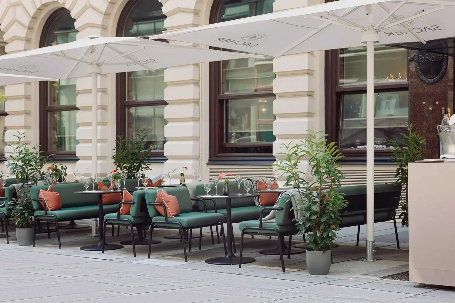 Ein leerer Außenbereich eines Restaurants mit grünen Sitzbänken und Tischen von Bene Outdoor, eingedeckt mit Besteck und Gläsern, unter großen weißen Sonnenschirmen, umgeben von Pflanzen und großen Fenstern an einem historischen Gebäude.