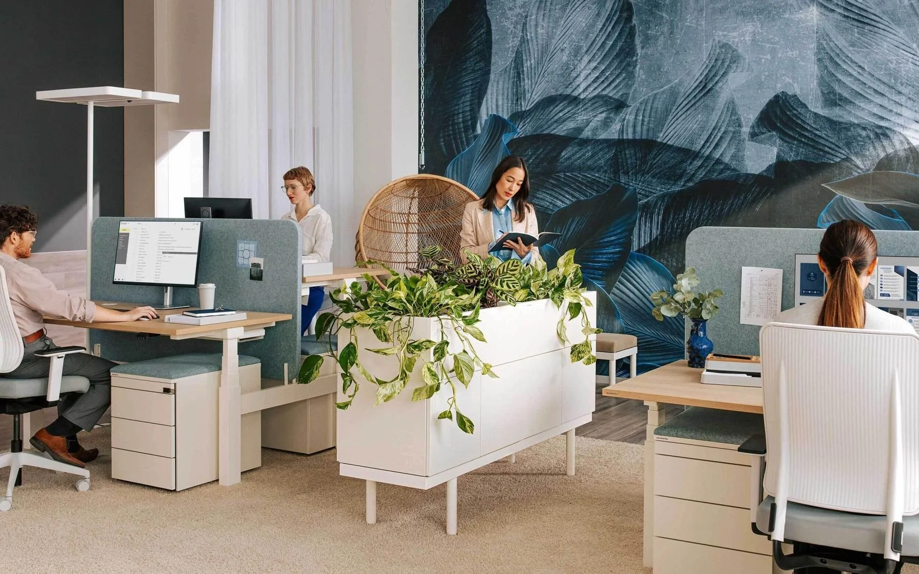 Moderner und freundlicher Büroarbeitsplatz von neudoerfler Büromöbel mit fünf Mitarbeiterinnen, Pflanzen, Wand mit Blättermuster, und hellem Ambiente. Raumwelten und Bürolösungen. Experte für Lern- und Arbeitswelten.