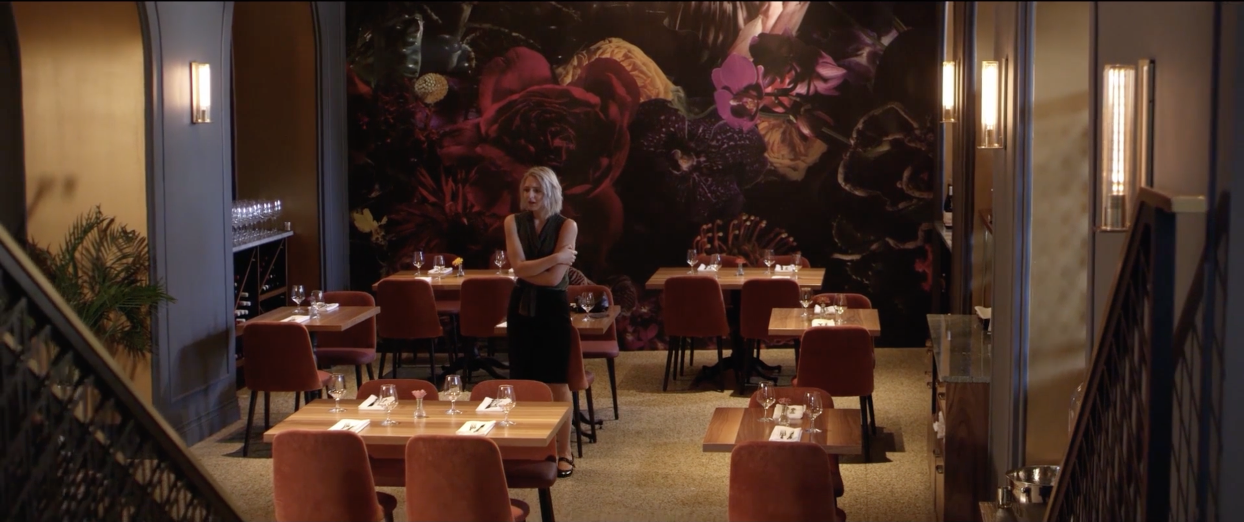 A woman with blonde hair in a black dress standing in a dimly lit restaurant with empty tables and glasses, a large floral mural on the back wall, and warm lighting.
