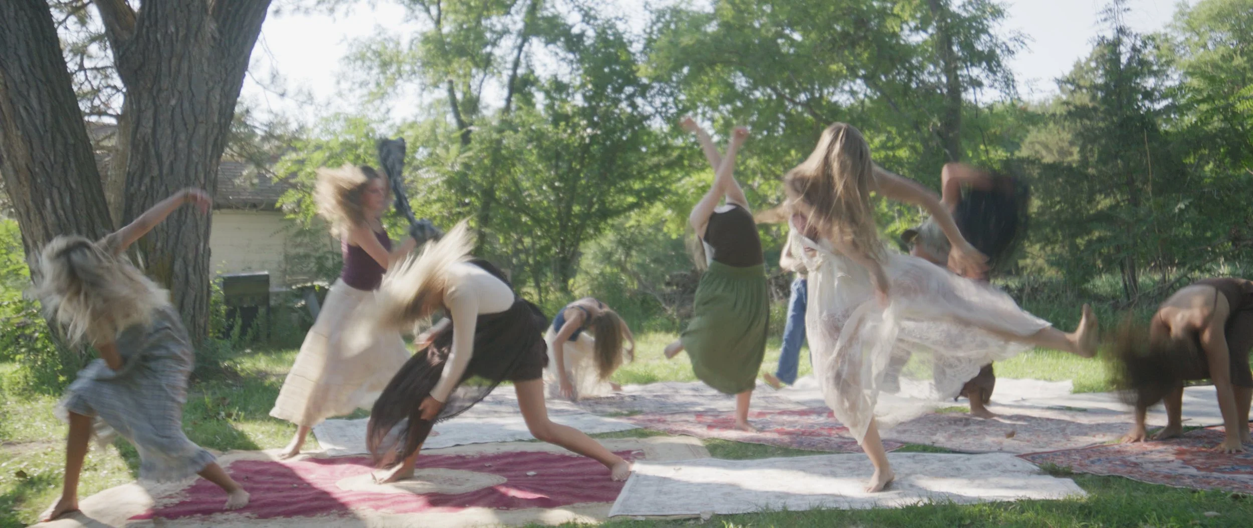 Several women and girls are dancing and spinning on rugs outdoors in a backyard with trees and greenery, with some of their hair and dresses flying due to movement.