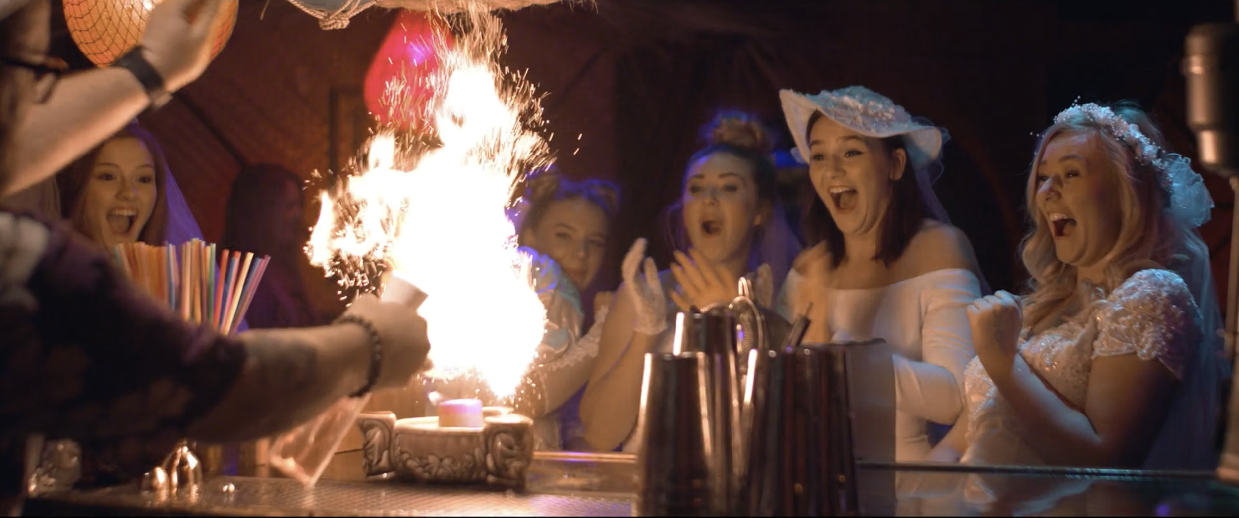 A group of women celebrating a birthday with a flaming cake and party hats, surrounded by drinks and party decorations.