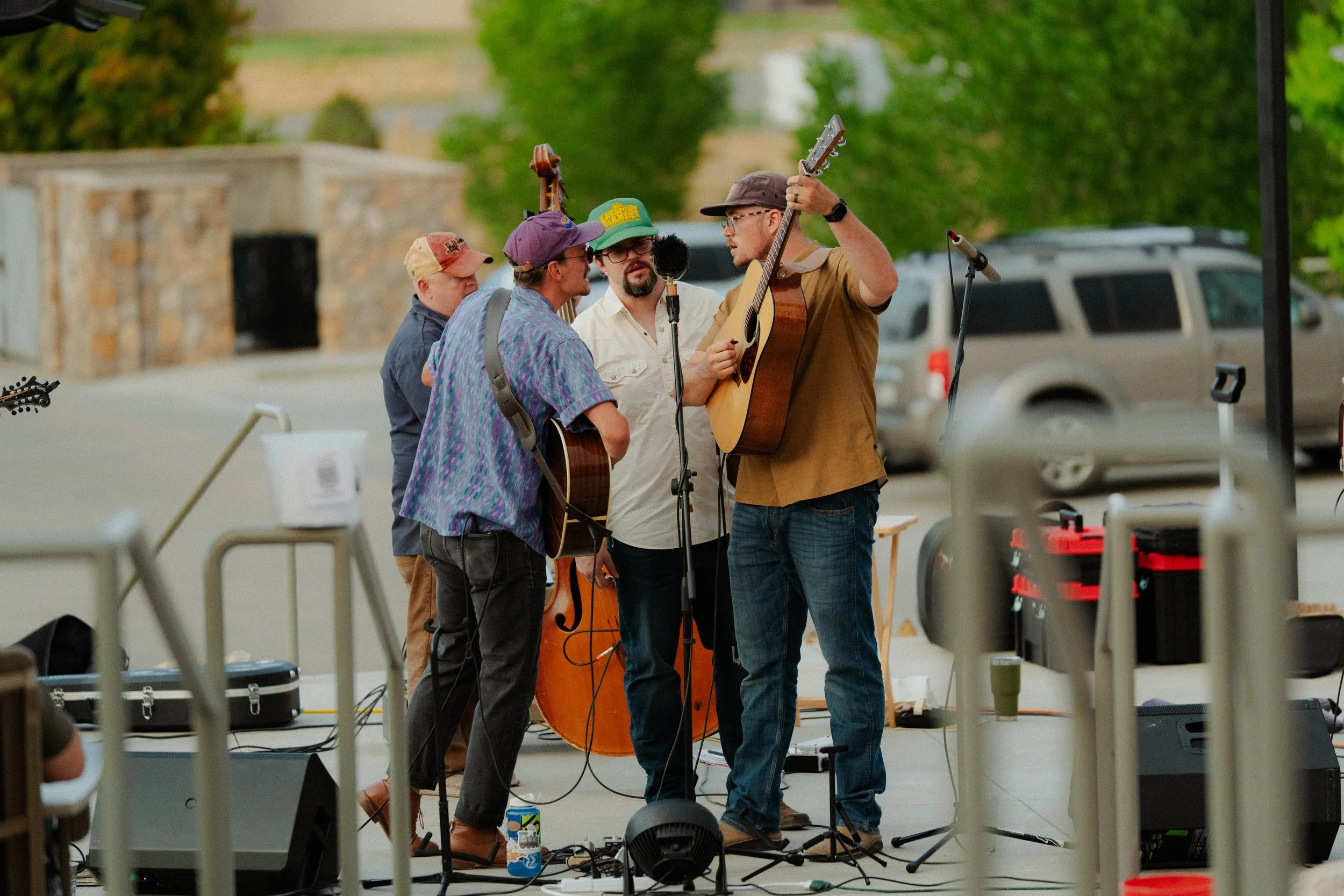 A group of five men on a stage, two with guitars, performing or rehearsing outdoors with a microphone, surrounded by equipment and trees in the background.
