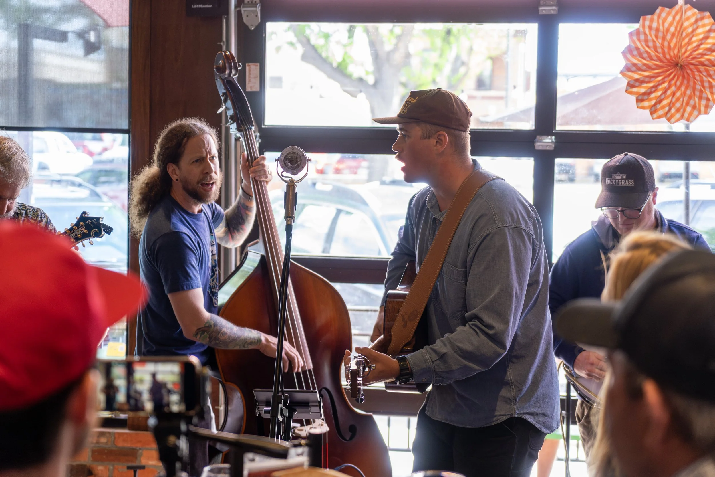 A group of musicians performing live indoors, with a man singing and playing a guitar, and another man playing a double bass, while people watch.