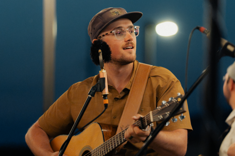 A man wearing glasses, a brown cap, and a brown shirt is playing an acoustic guitar and singing into a microphone.