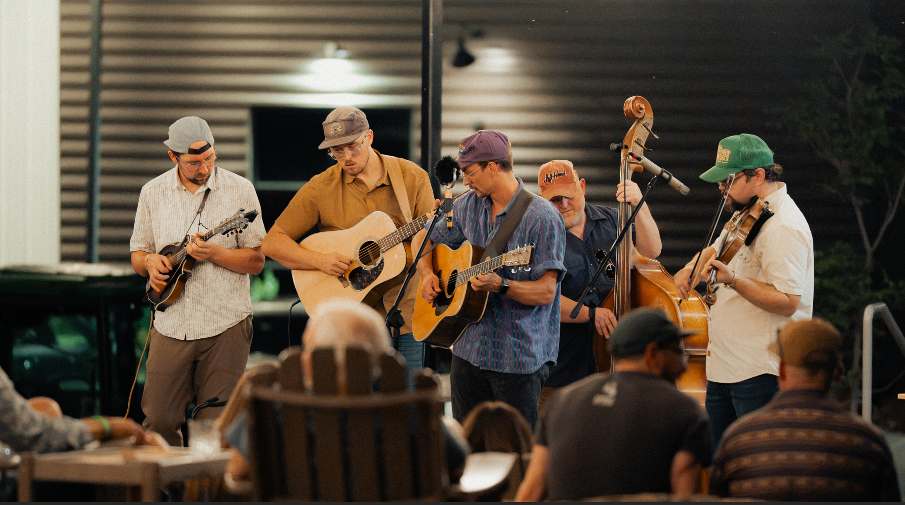 A group of five musicians playing musical instruments on a stage with an audience watching. The musicians include two with acoustic guitars, one with a mandolin, one with a double bass, and one with a violin. They are performing in a casual setting outdoors at night.