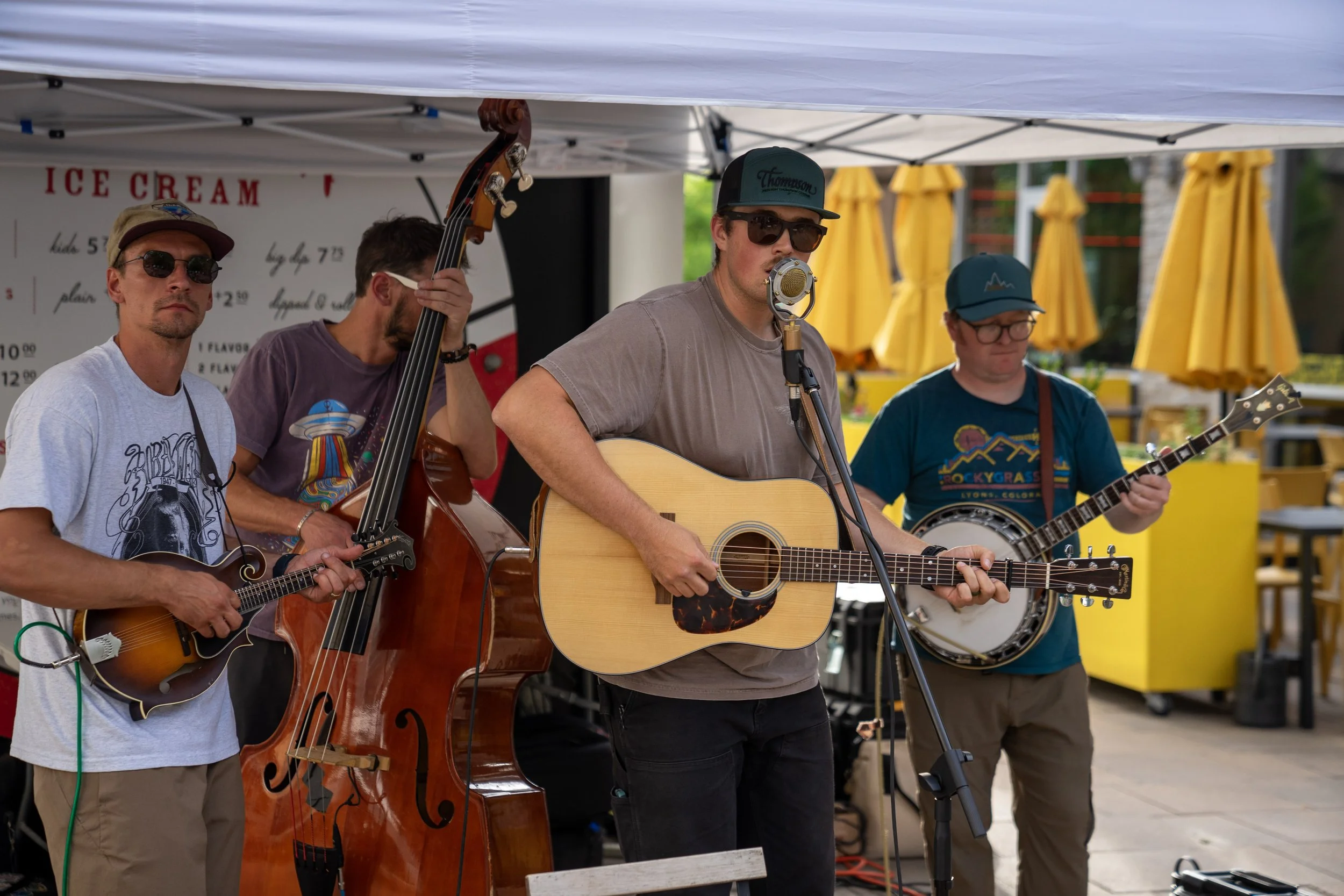 A group of four men playing musical instruments at an outdoor event. One is playing a guitar and singing into a vintage microphone, another is playing a banjo, the third is playing an upright bass, and the fourth is playing a guitar. They are under a