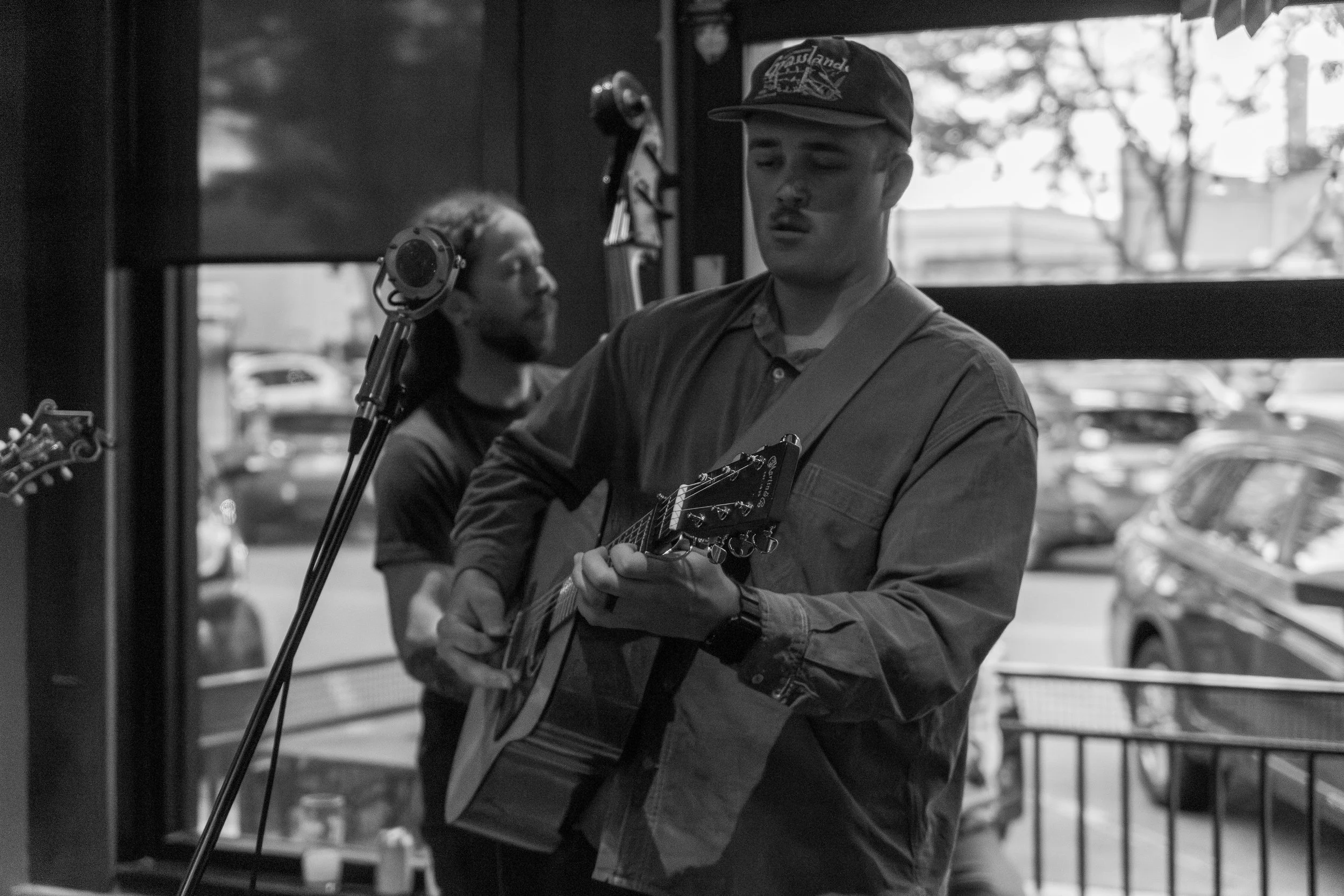 A man playing an acoustic guitar in a cafe or shop, wearing a baseball cap and a casual shirt, with another person in the background, possibly singing or listening.