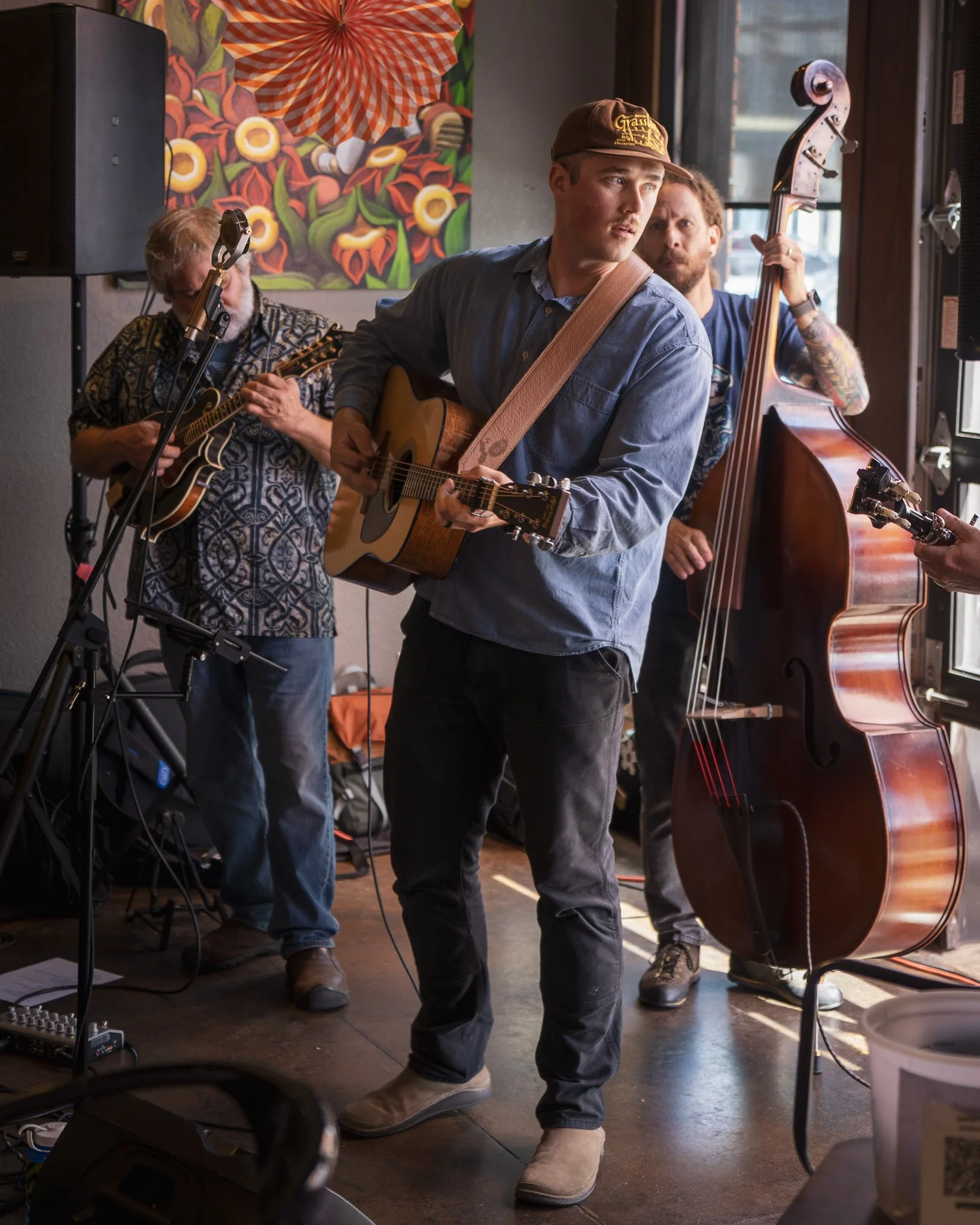 Three musicians performing inside a cafe or record store with colorful artwork on the wall; the man in front is playing acoustic guitar, wearing a brown cap, blue shirt, and tan boots; the man behind him is playing an upright bass; the third man to t