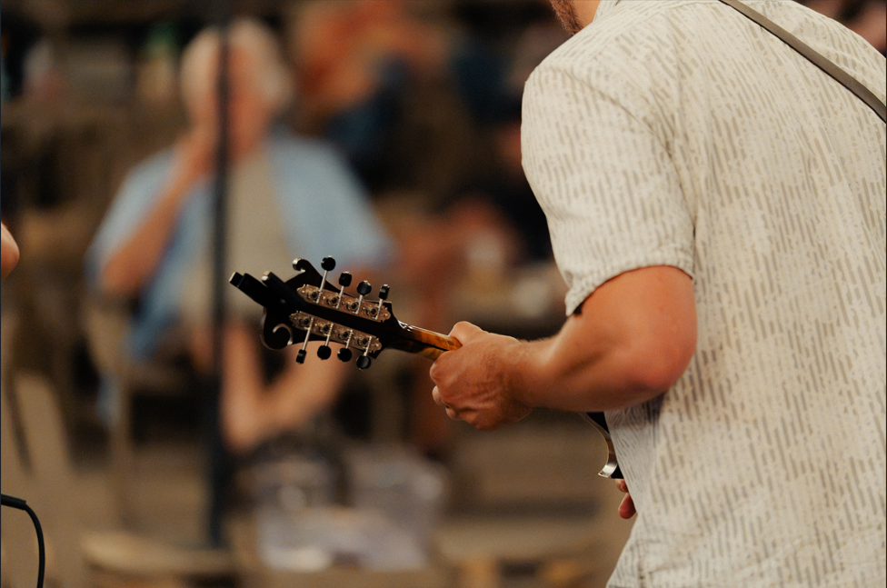 A man playing a guitar outdoors with a blurred crowd in the background.
