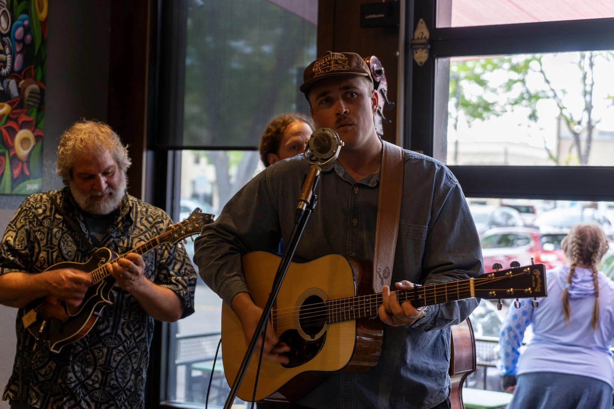 Two young men playing guitars and singing into a microphone in a cozy indoor setting with natural light coming through large windows, dressed casually and focused on their music.