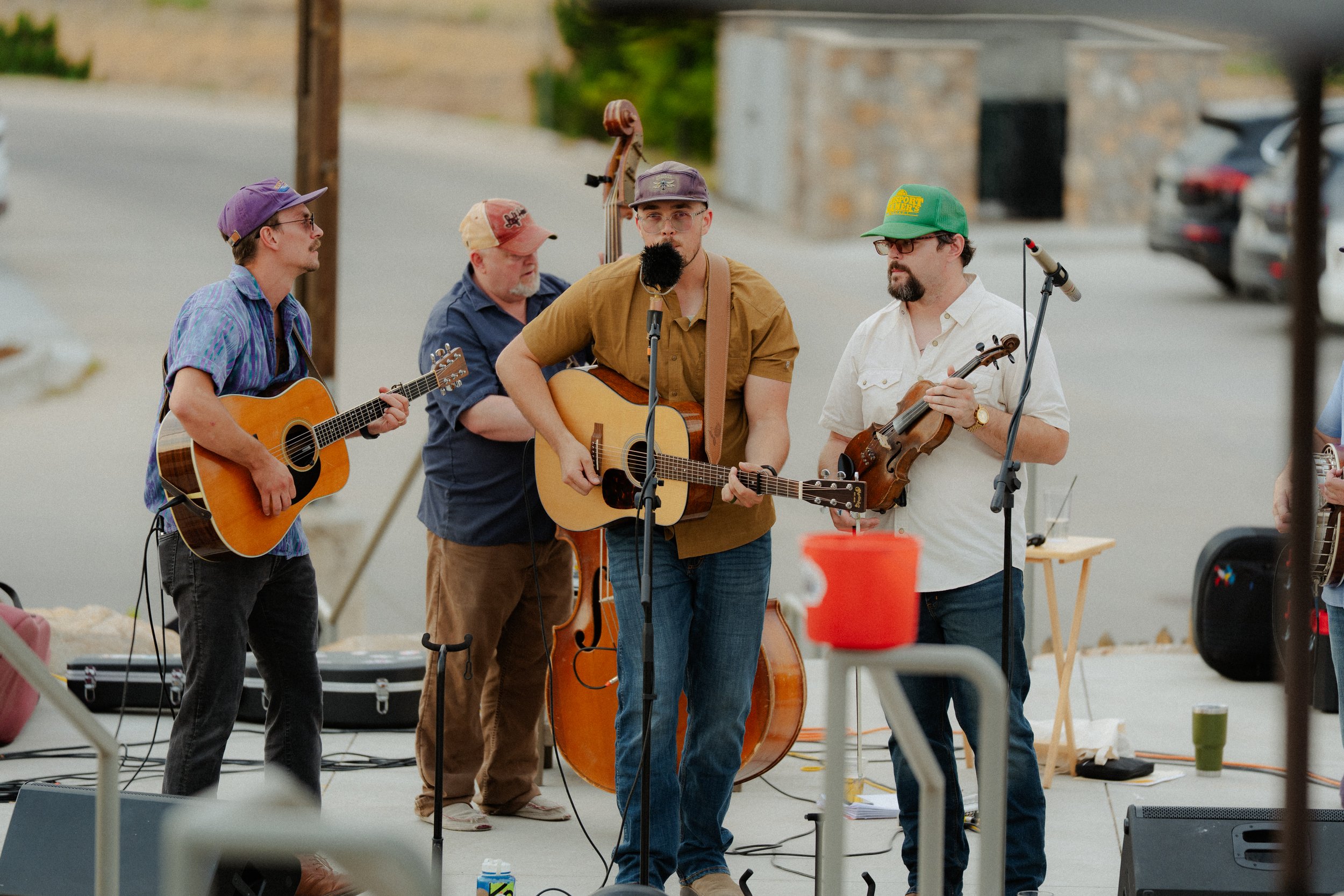A group of four male musicians is performing outdoors, playing guitars and a violin, with microphones in front of them. They are casually dressed and wearing hats, with a background of parked cars and a stone building.