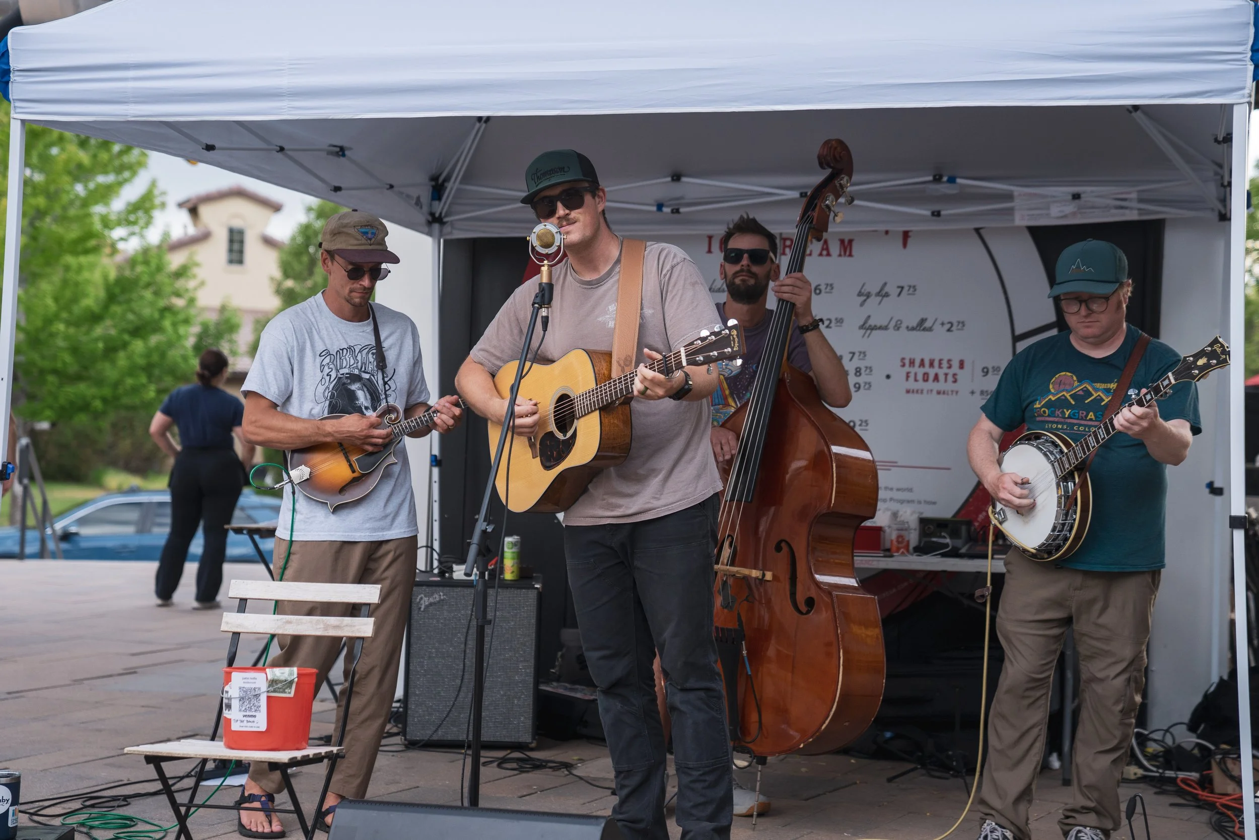A band of four men performing live music under a white canopy tent outdoors. One is playing a mandolin, another an acoustic guitar and singing into a microphone, another upright bass, and the fourth also playing a guitar. They are wearing sunglasses 