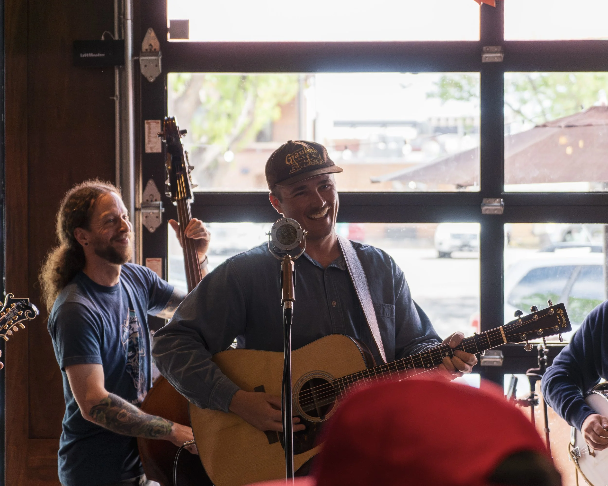 Two musicians performing indoors, one playing an acoustic guitar and the other holding a stringed instrument, with a window showing trees and buildings outside.