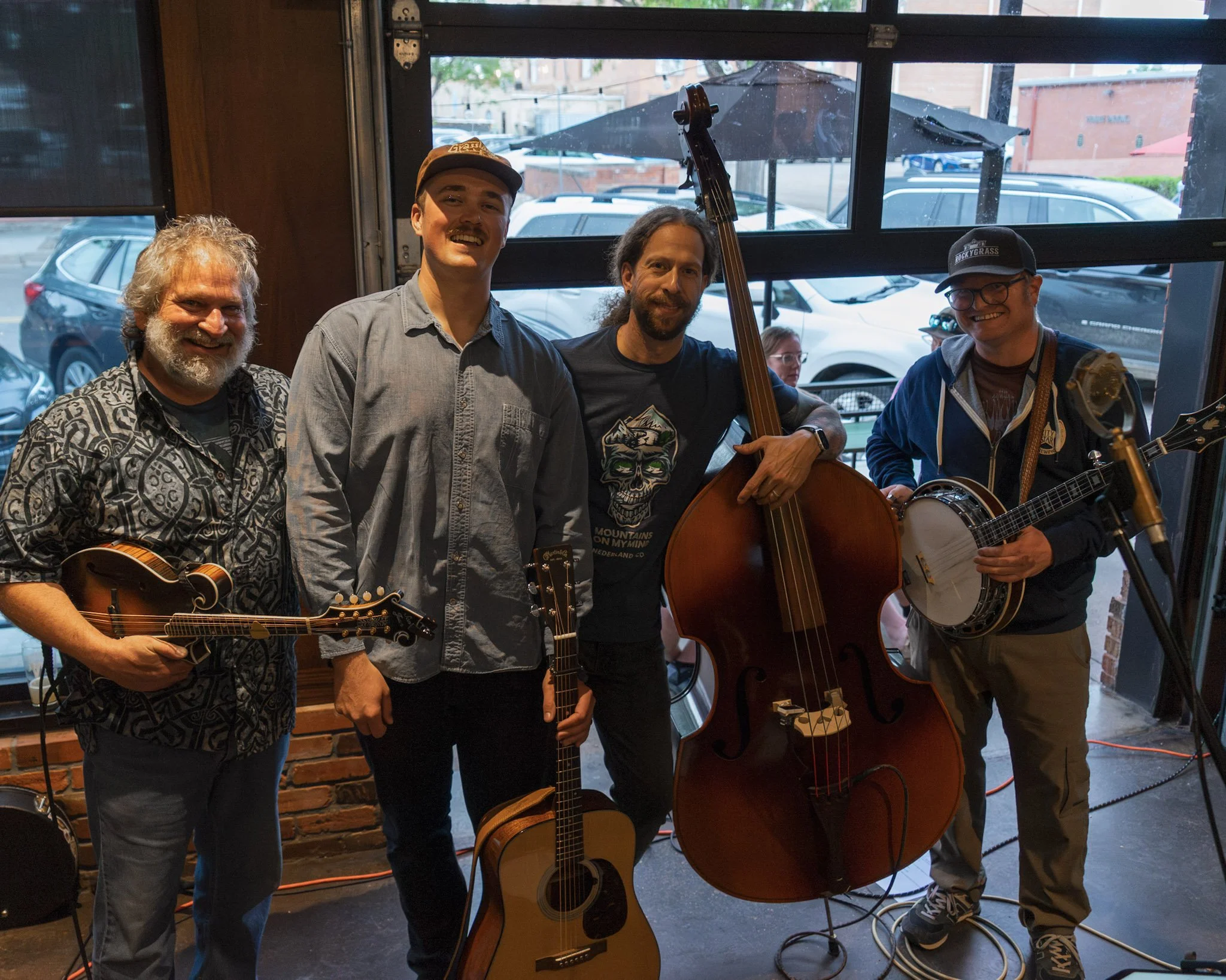 Four men standing inside a restaurant with guitars, two of them are wearing hats, one is holding a double bass, and another is smiling at the camera. Outside the window, parked cars and an umbrella are visible.