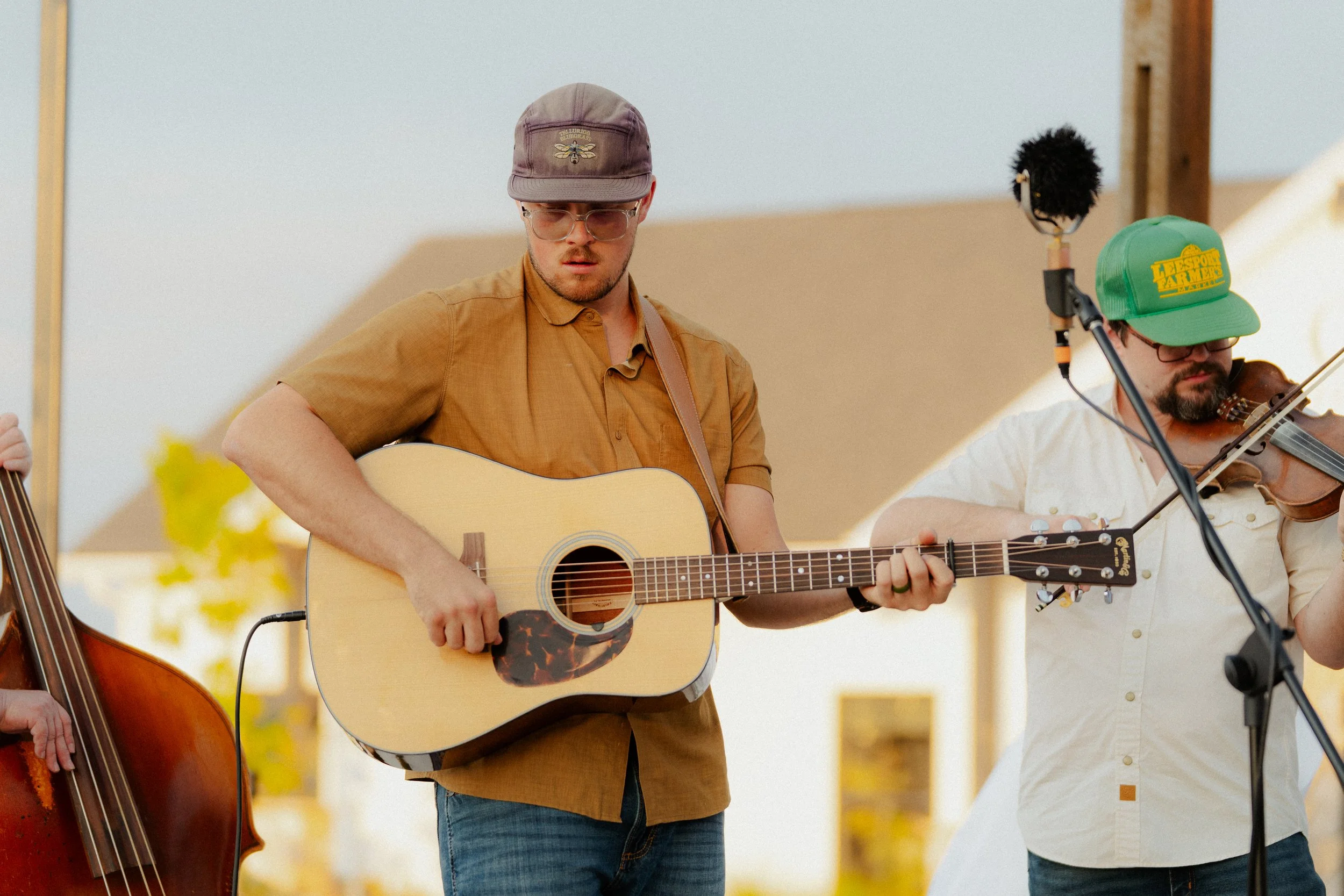 A man wearing glasses and a gray cap is playing an acoustic guitar on stage. He is dressed in a brown shirt and blue jeans. Behind him, another musician wearing a green baseball cap and glasses is playing a violin. The background shows a clear sky an