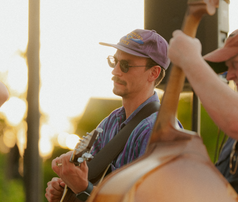 A young man wearing a purple cap, sunglasses, and a plaid shirt playing an acoustic guitar outdoors at sunset.