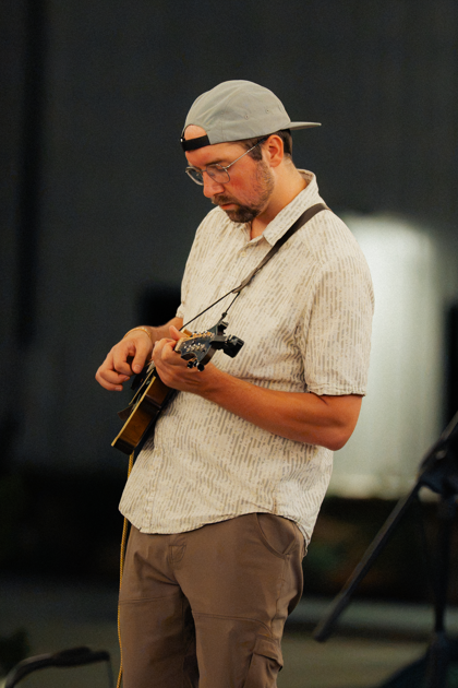 A man wearing glasses, a gray cap, and a beige short-sleeve shirt playing an acoustic guitar in an indoor setting.