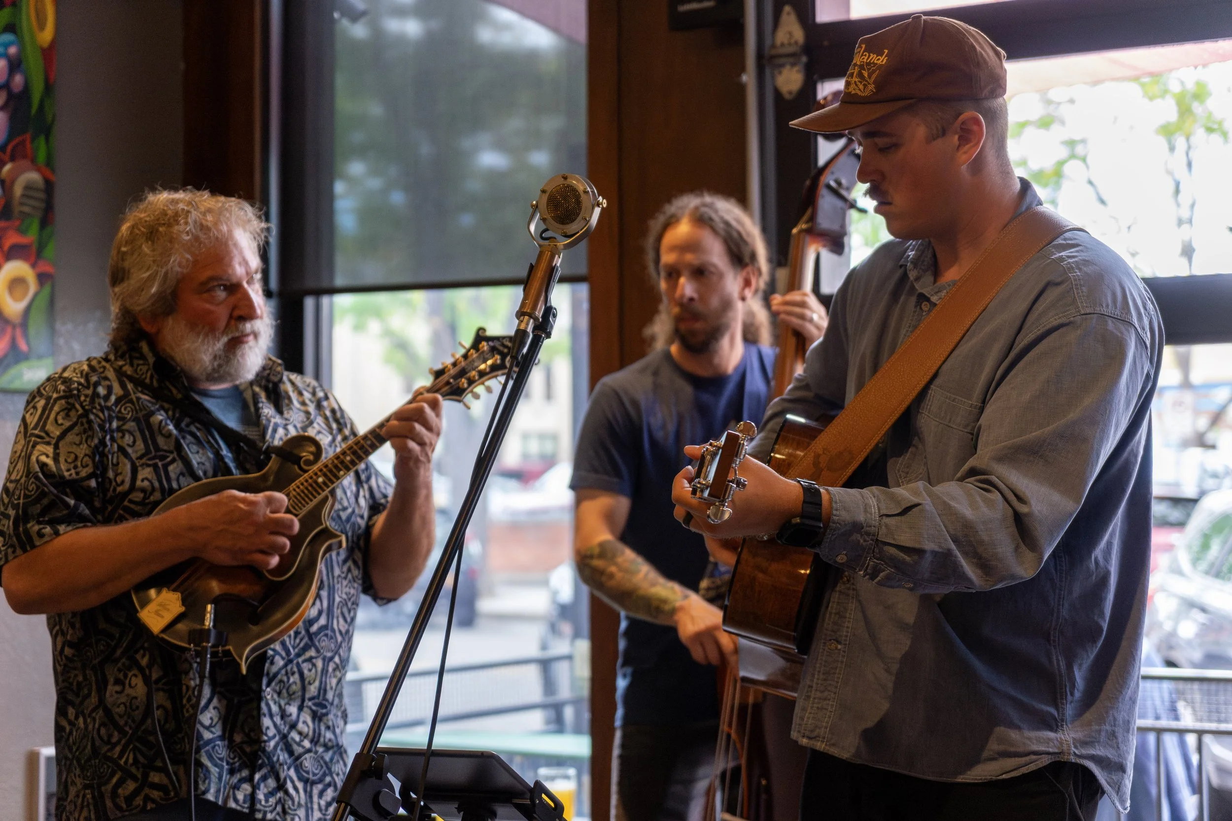 Three musicians playing instruments inside a room with large windows, natural light, and colorful artwork on the wall.