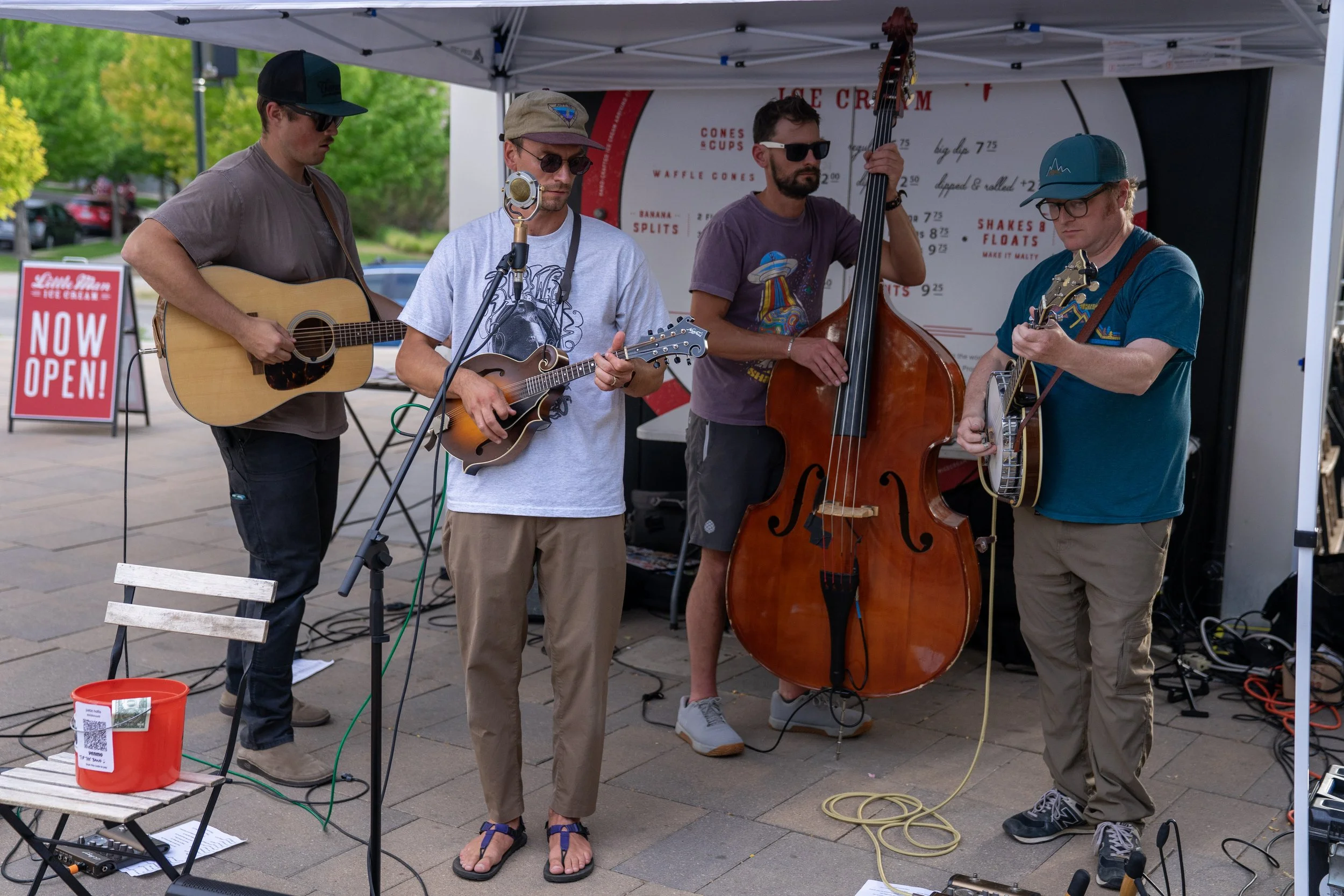 Four musicians performing with guitars, mandolin, and upright bass under a canopy at an outdoor event, with a menu board and a sign reading 'Now Open!' in the background.